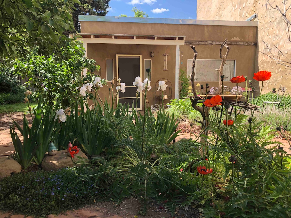 A tranquil garden scene features a variety of blooming flowers, including striking red poppies and elegant irises, surrounding a cozy cottage. The outdoor space is characterized by lush greenery and a pathway leading to the inviting entrance of the cottage, beneath a clear blue sky.