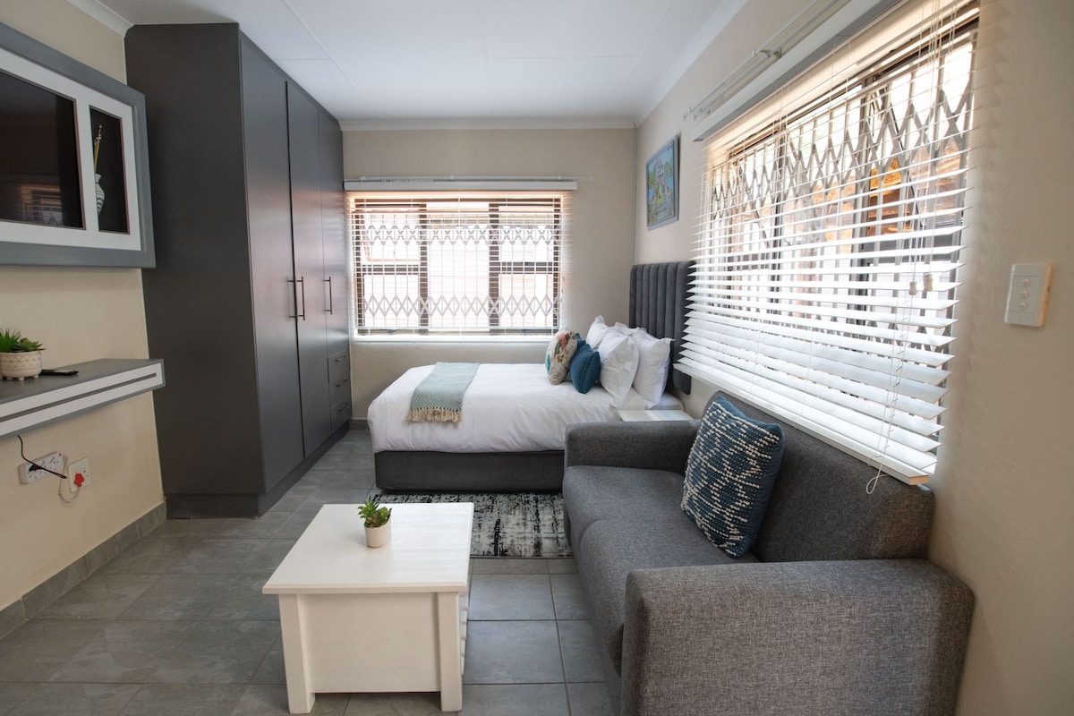 A compact living space is highlighted by a cozy seating area featuring a gray sofa with decorative pillows. A spacious bed is positioned against the wall, adorned with white linens and accent pillows. Large windows with blinds allow natural light to fill the room.