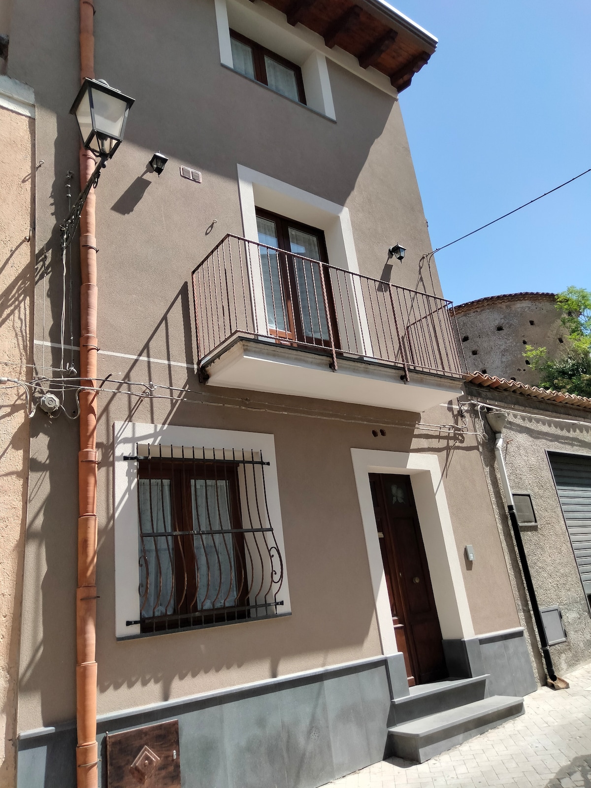 A modern building exterior features a clean façade with light-colored walls and wooden accents. A small balcony is present on the second floor, with decorative iron railings framing the windows. The entrance door is framed by a subtle architectural detail, adding character to the overall design.