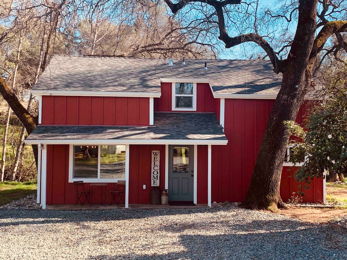 The bright red farmhouse exterior is framed by large trees, creating a picturesque setting. Two outdoor chairs are positioned by the entryway, inviting guests to relax. A welcoming sign hangs near the door, contributing to the home's friendly atmosphere.
