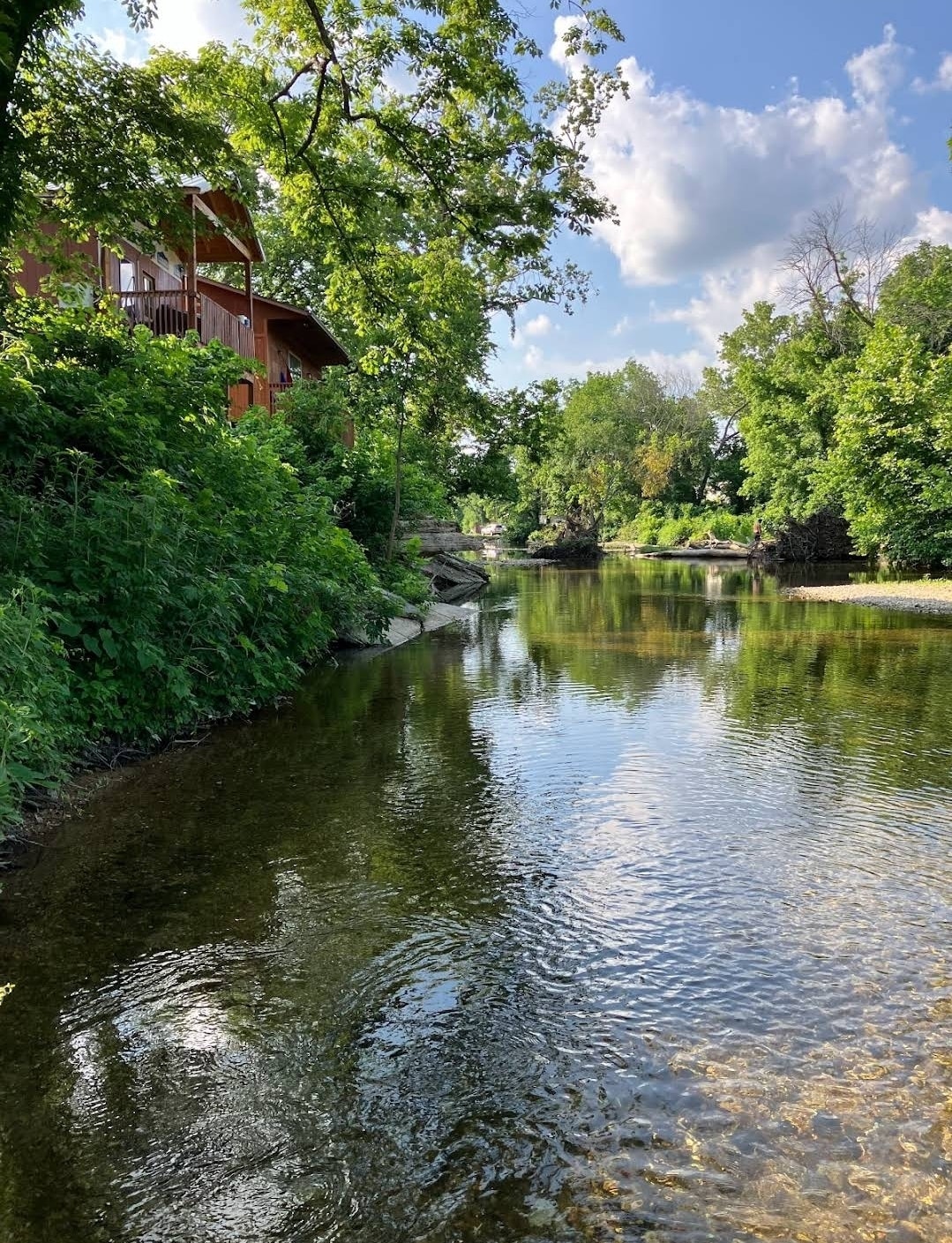 The image captures a serene waterway flowing gently beside a cabin surrounded by lush greenery. Reflections of trees and a bright sky are visible on the calm surface of the water, which varies in depth near the rocky banks.