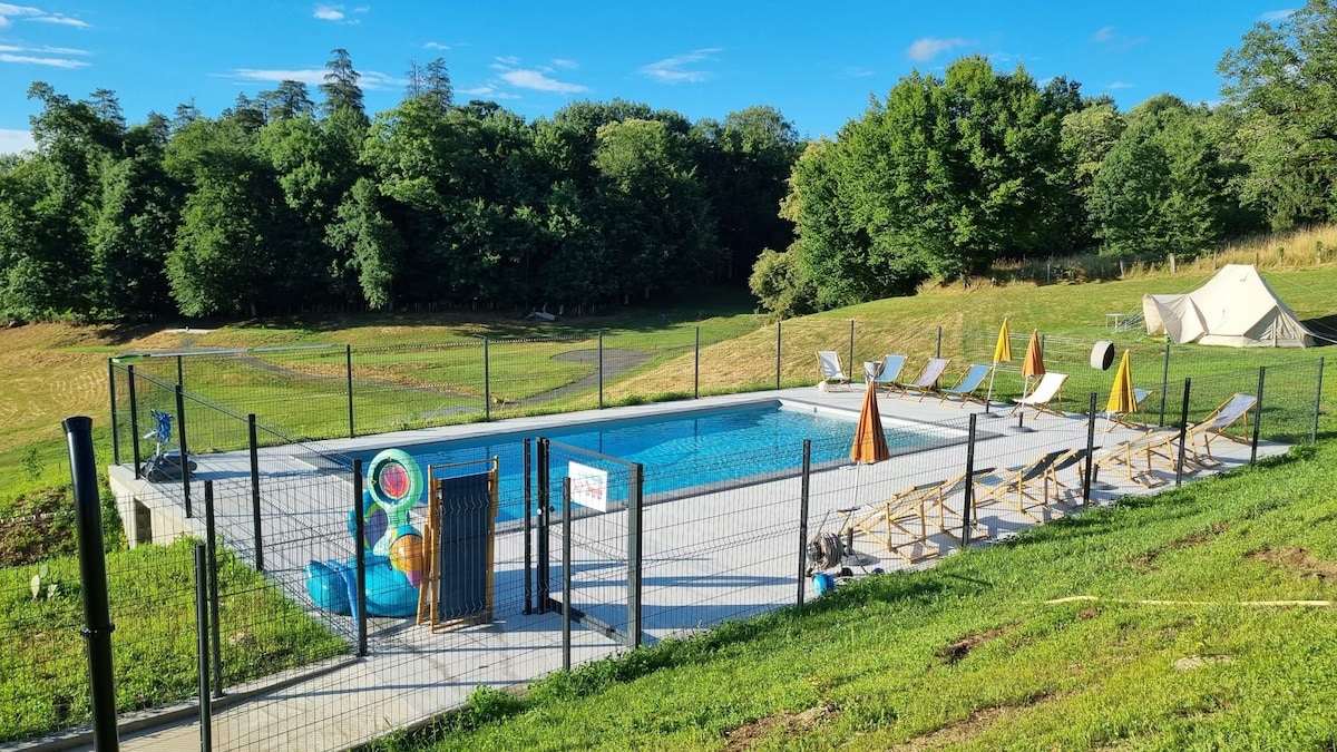 An outdoor salt-water pool is surrounded by an enclosed area, featuring several lounge chairs positioned for relaxation. Sun umbrellas provide shade, while a grassy area and trees create a natural backdrop. A tent can be seen in the distance, contributing to the outdoor ambiance.