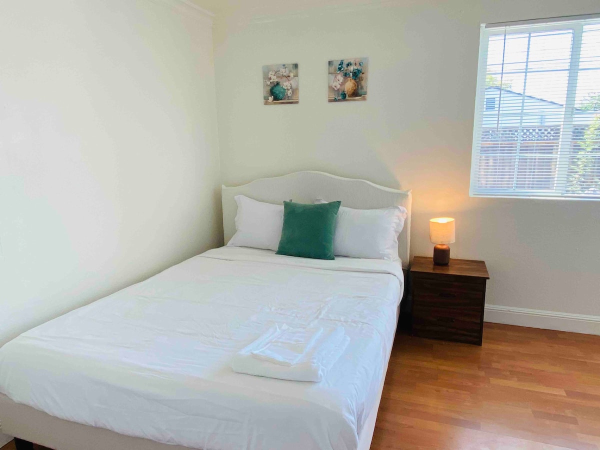 A bright bedroom features a spacious bed with white linens and a green accent pillow. A nightstand holds a soft-glowing lamp, and two framed artworks adorn the walls. Natural light enters through a window with a view of the outdoors.
