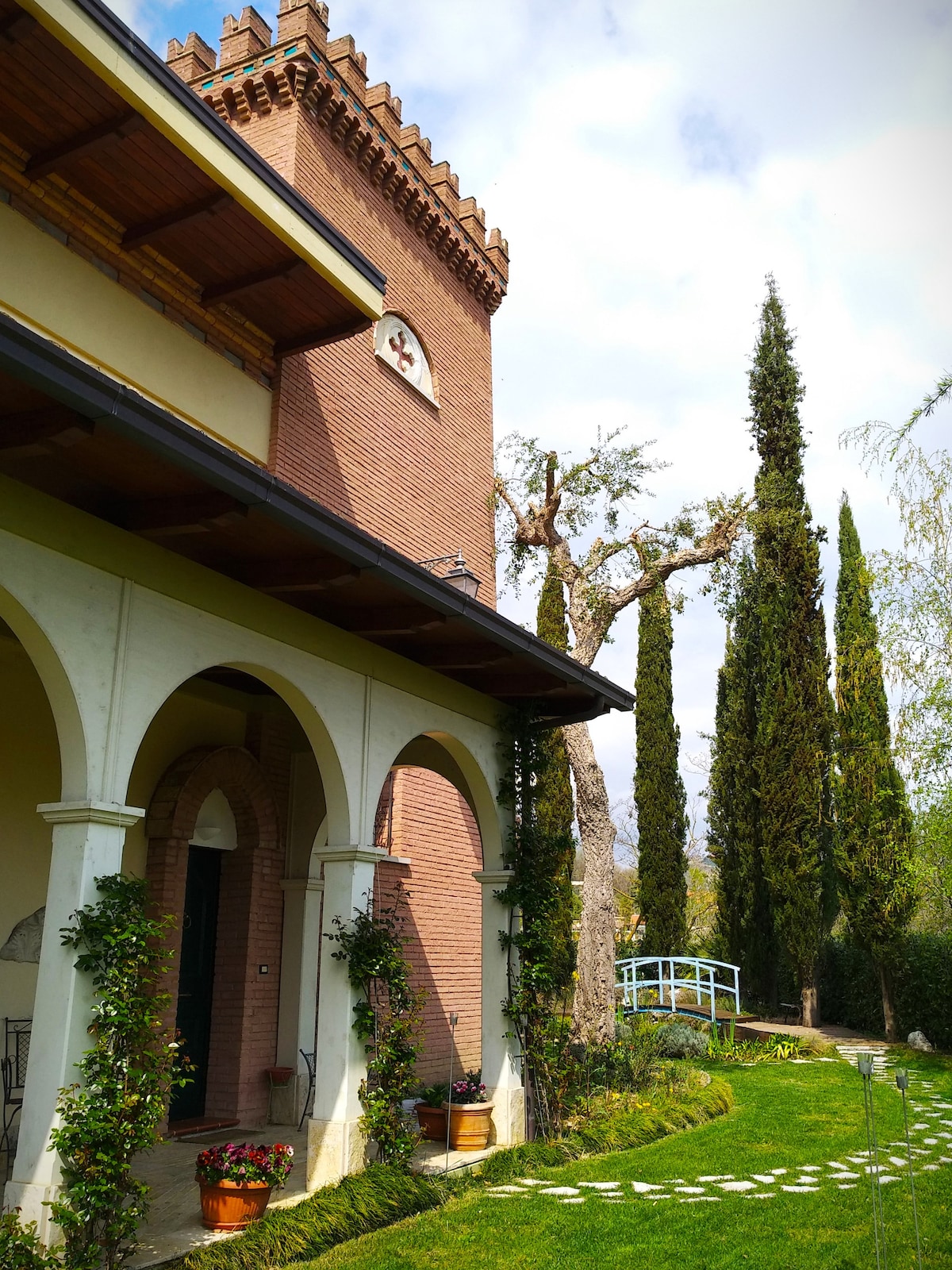 The image depicts a charming architectural structure featuring arched doorways and a decorative tower. Surrounding greenery includes tall cypress trees and a manicured lawn with a stone pathway leading to a wooden bridge. Flower pots enhance the entrance with a touch of color.