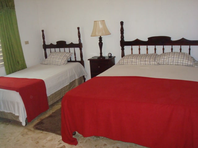 Two neatly made beds are positioned in a well-lit room, featuring simple wooden headboards. A bedside table with a lamp sits beside one bed. Red blankets are draped over both beds, contrasting with the white linens and creating a cozy look.