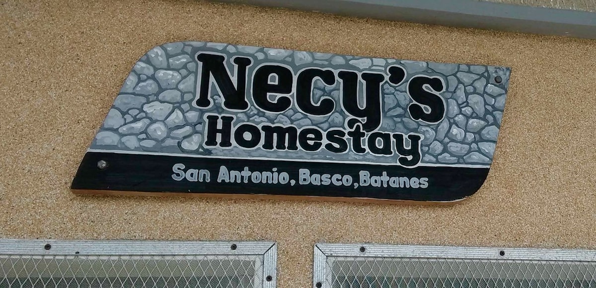 A stone sign is displayed, reading 'Necy’s Homestay' in bold black letters against a textured background. Below, 'San Antonio, Basco, Batanes' is written in white. The design highlights the homestay's welcoming identity.