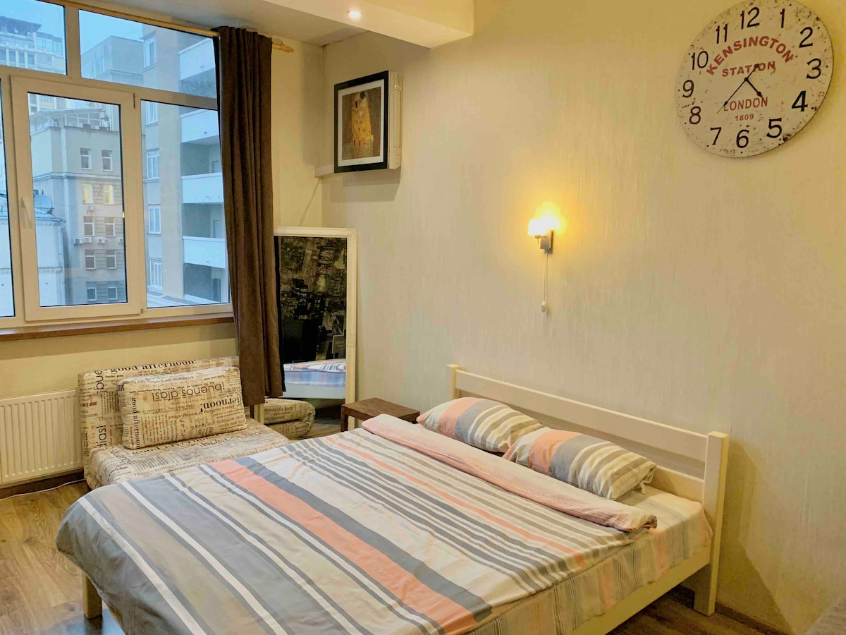 A bright sleeping area features a spacious bed dressed with striped linens, complemented by a soft sofa nearby. Natural light enters through the large window, while a wall clock and framed artwork add subtle details. The room's design is contemporary and inviting.