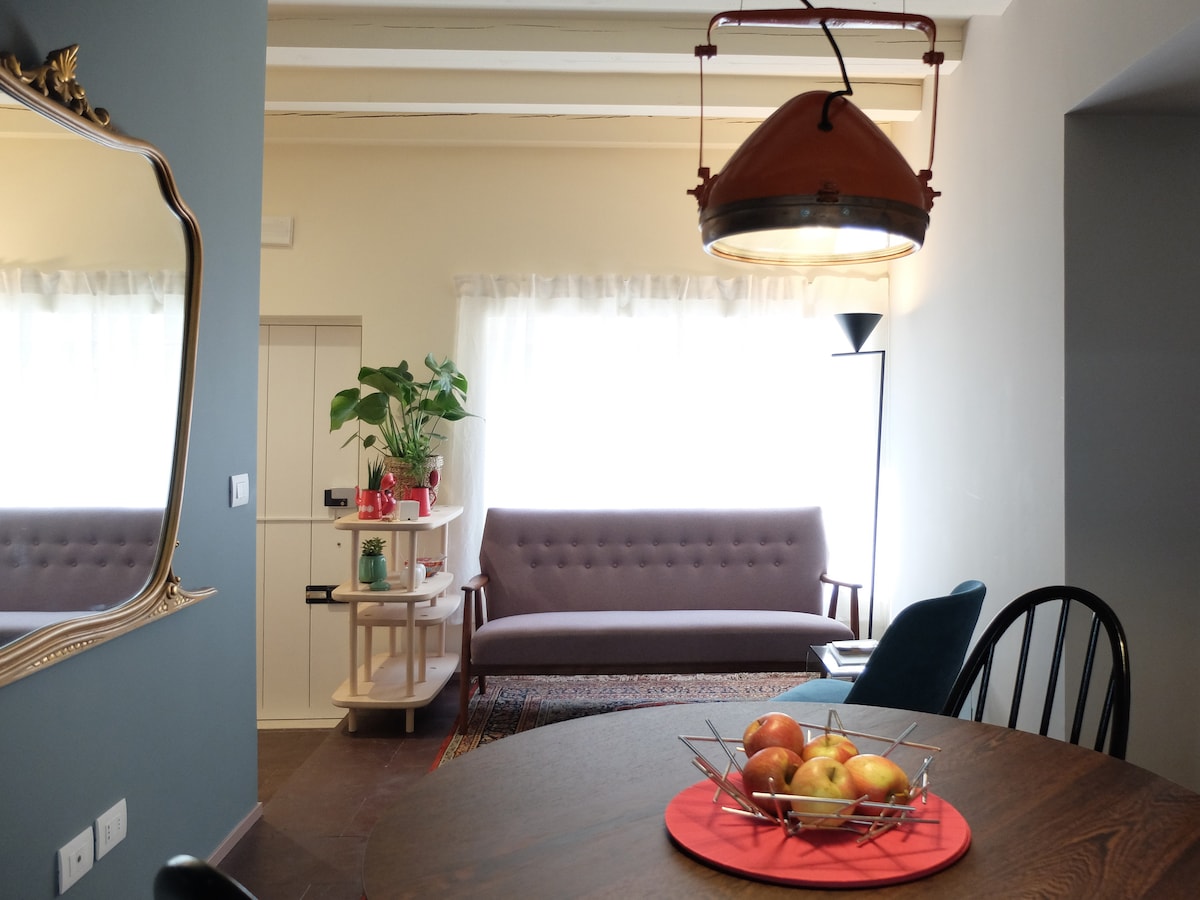 A living area showcases a purple sofa under soft natural light from a large window. A wooden dining table with black chairs sits in the foreground, adorned with a round red placemat holding a bowl of apples. A floor lamp and greenery add a touch of vibrancy.