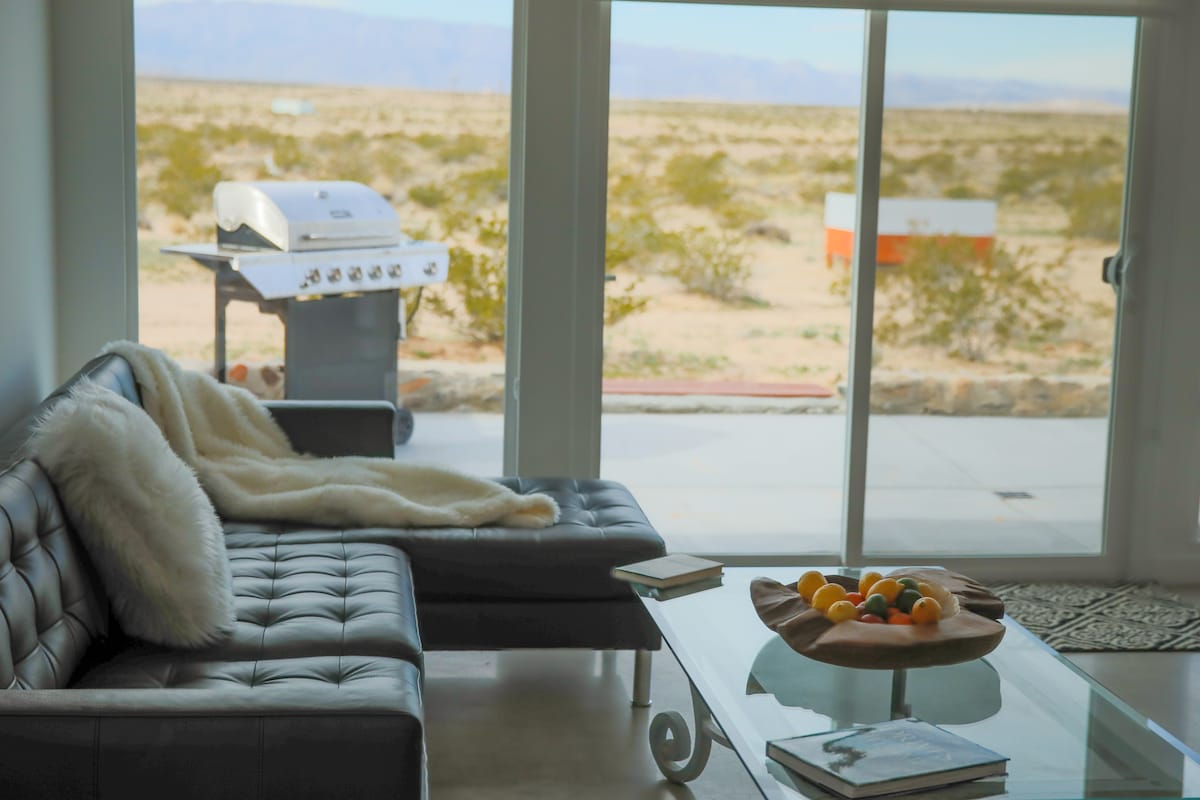 A modern living area features a sleek sectional sofa draped with a soft throw. A glass coffee table holds a bowl of fresh fruit. Large sliding doors provide a view of the desert landscape, with a barbecue grill visible on the patio.