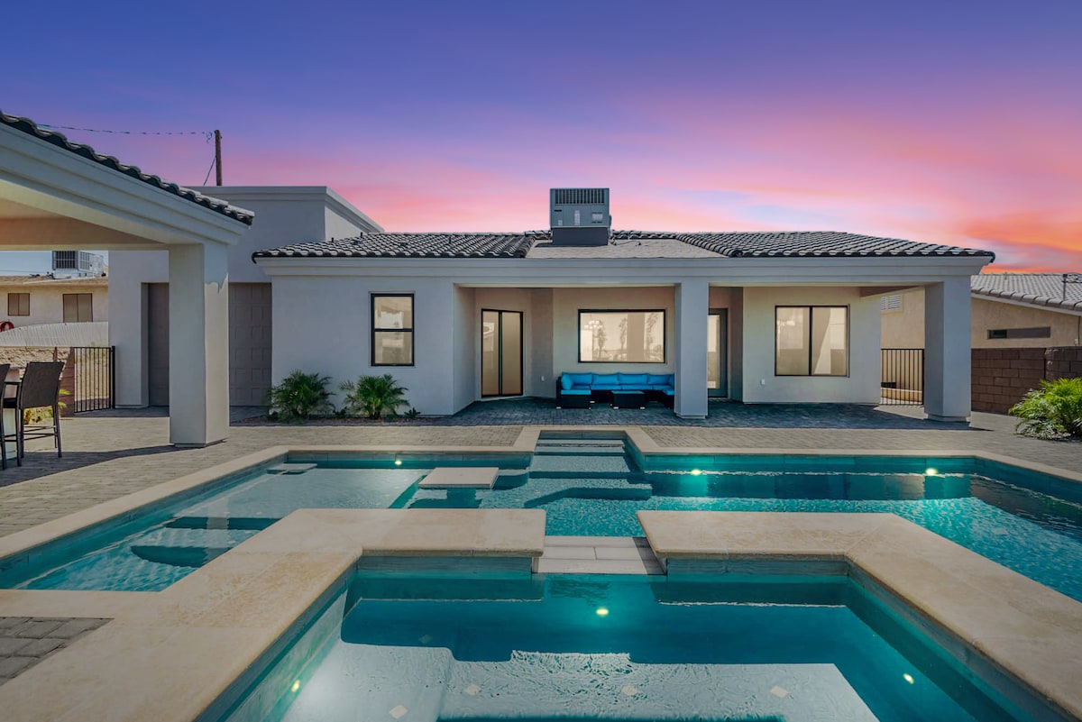 A modern home exterior is visible against a vibrant sunset sky. The inviting pool features a spa and is surrounded by a paved patio area. Large windows reveal portions of the interior, and outdoor seating is arranged nearby, enhancing the space for relaxation and entertainment.