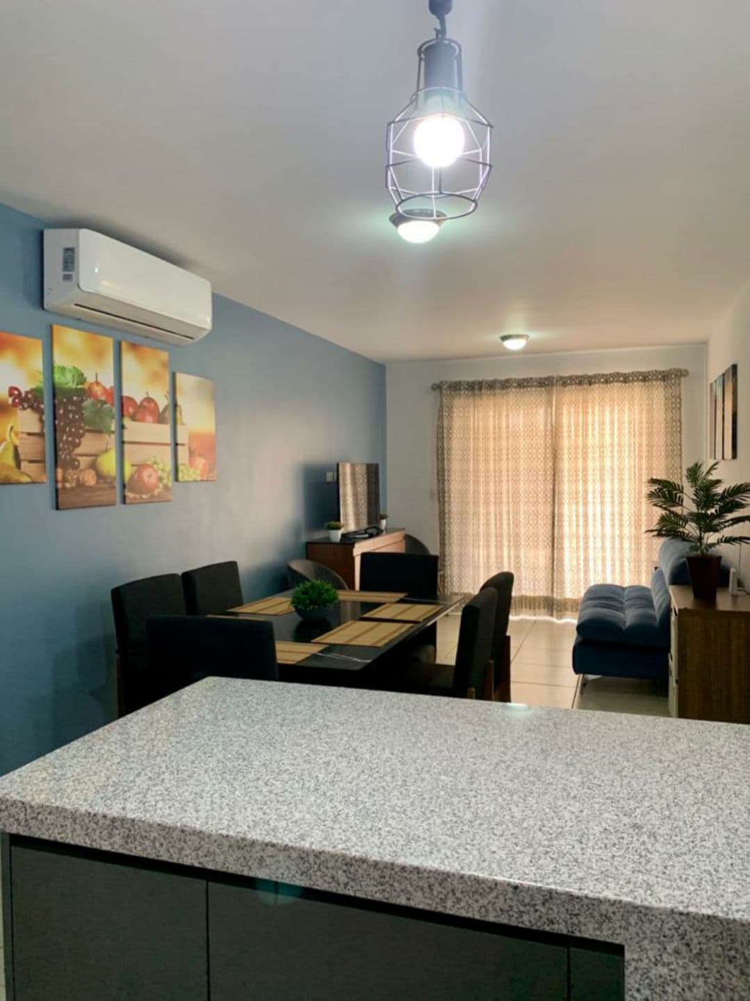 A modern living space features a light-colored granite countertop, with a dining area showcasing a table surrounded by dark chairs. A comfortable sofa is positioned nearby, facing windows dressed in light curtains. Artwork depicting fruits accents the blue walls.