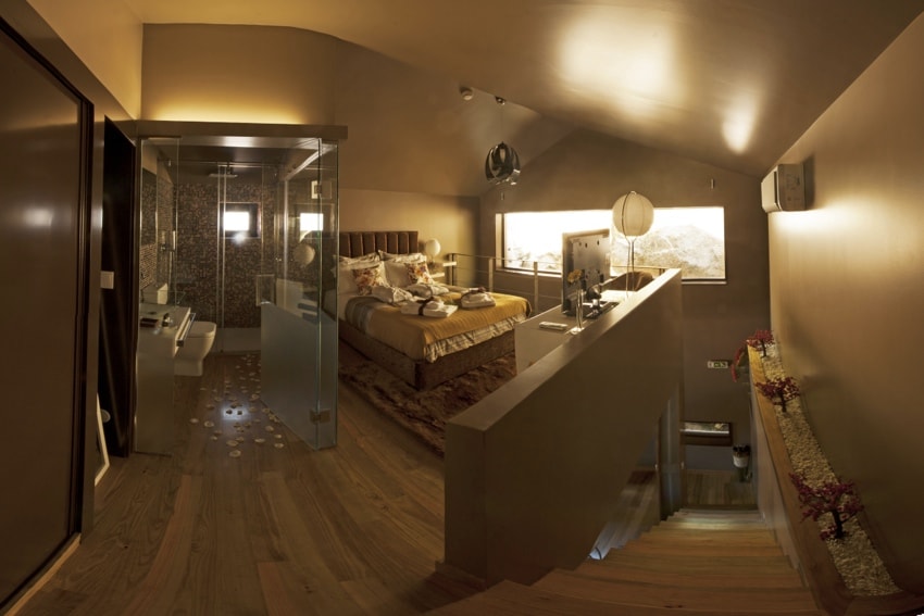 An open-concept bedroom showcases a spacious sleeping area with a comfortable bed adorned with soft bedding. Natural light filters through large glass windows, illuminating the room. A glass partition leads to the modern bathroom, while wooden flooring adds warmth to the ambiance.