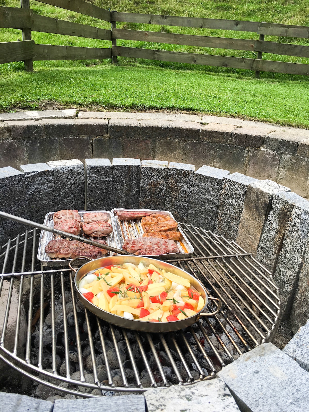 A stone fire pit is featured, with a grilling rack holding various meats and a pot of vegetables steaming above the flames. The grassy area surrounding the pit is enclosed by a wooden fence, creating a private and inviting space for outdoor gatherings.
