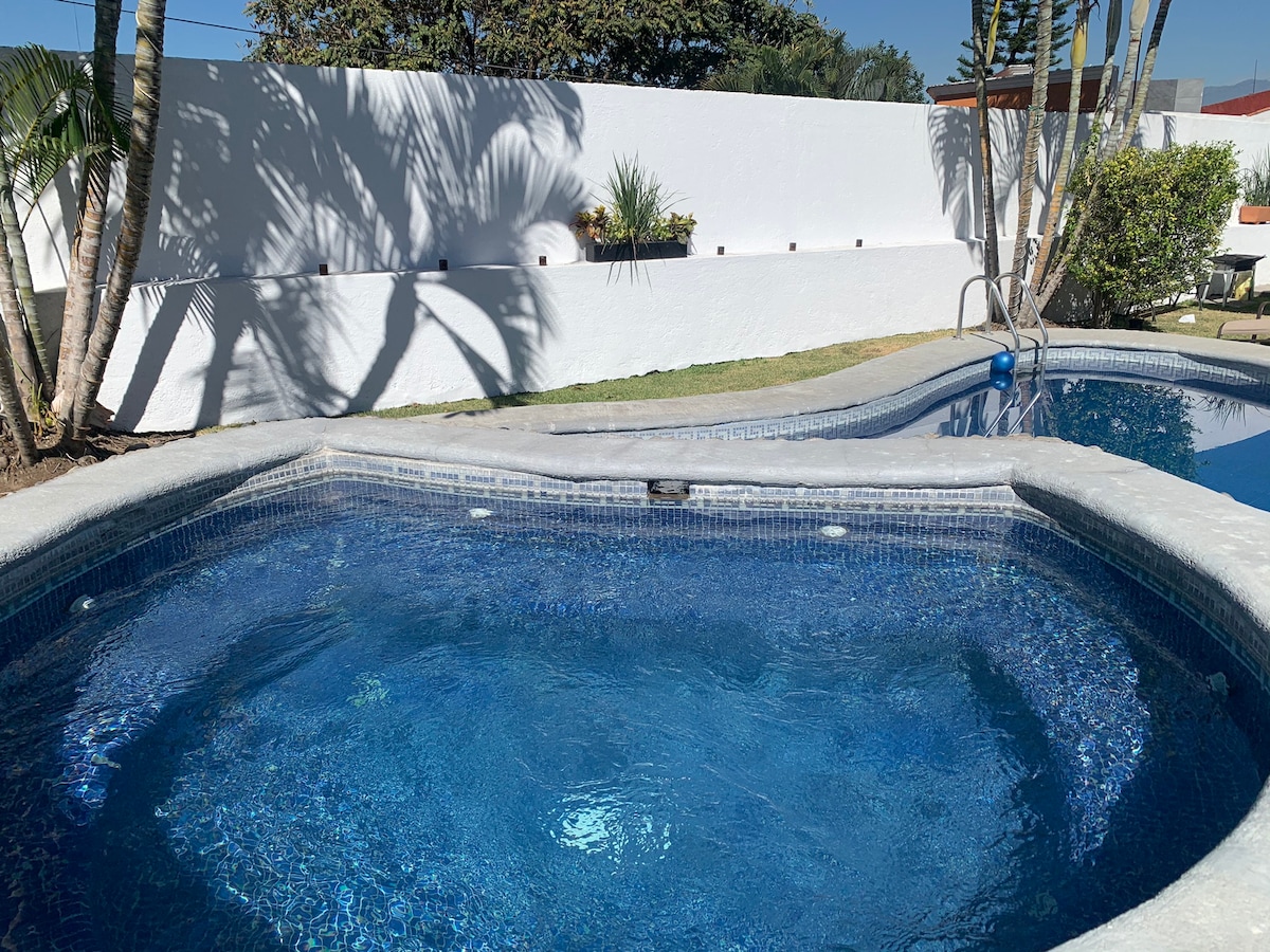 A heated jacuzzi is surrounded by a tiled border, inviting relaxation. The clear blue water reflects sunlight, while palm leaves cast gentle shadows. Nearby, a larger pool can be seen, enhancing the outdoor space of the garden.
