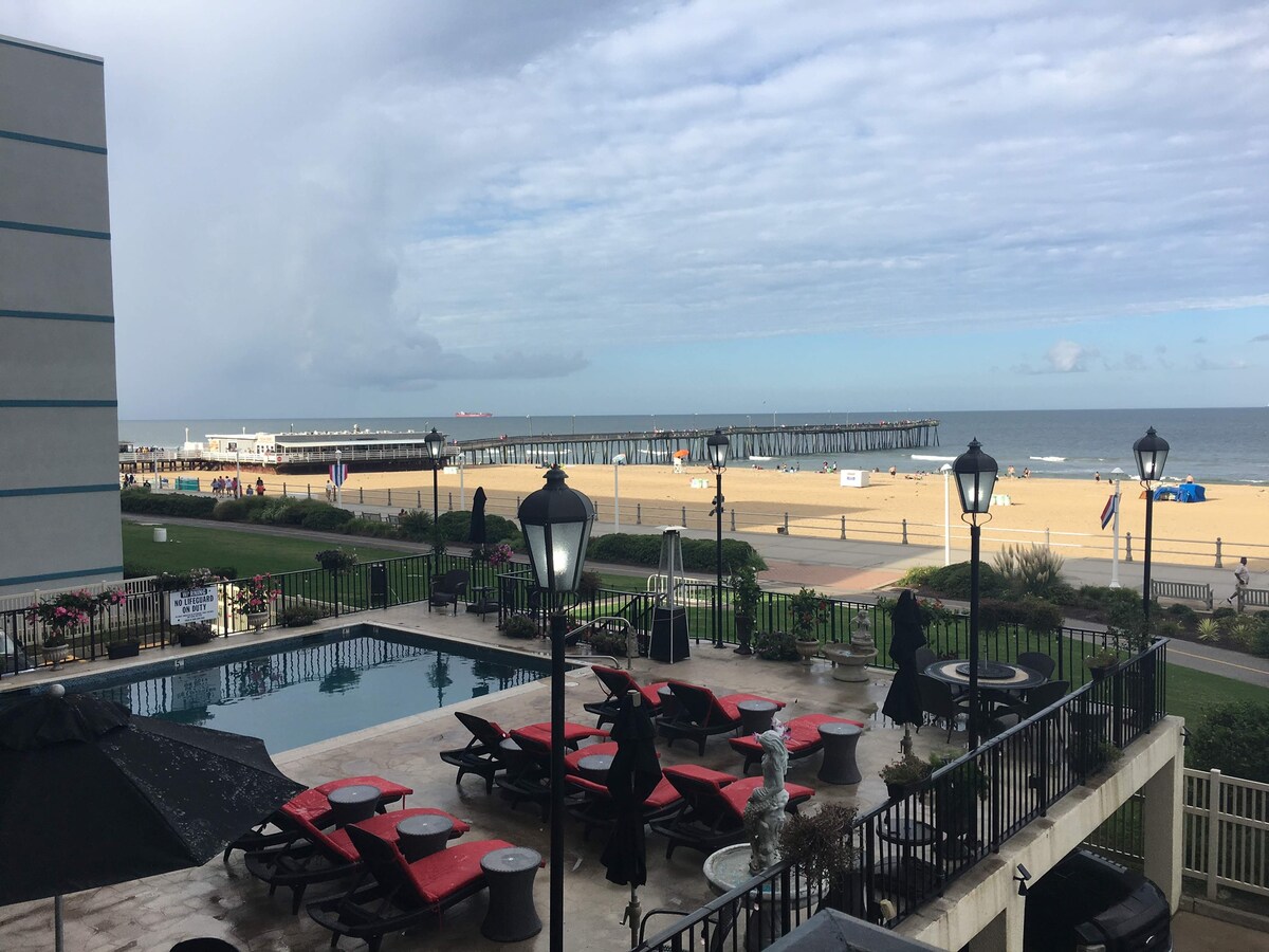 A view overlooking a beach area features a sandy shoreline and a fishing pier in the distance. The image includes a pool area surrounded by lounge chairs and umbrellas, with decorative lampposts providing ambient lighting.