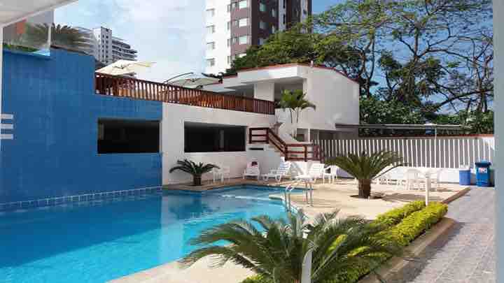 The image displays a communal outdoor pool area surrounded by palm trees and lounge chairs. Clear water reflects the blue sky, while a modern building offers shaded seating above. Tropical landscaping enhances the tranquil setting, creating a welcoming atmosphere for relaxation.