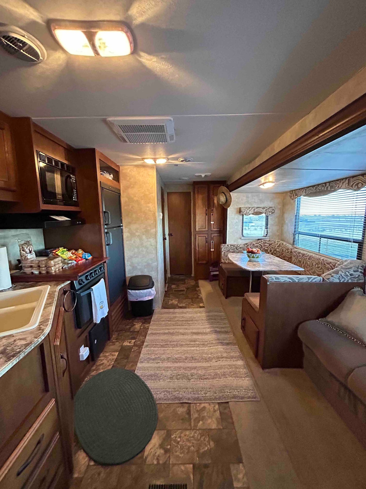 A cozy interior of a fully furnished RV is displayed, featuring wooden cabinetry, a compact kitchen area with essential appliances, and a comfortable seating space. Natural light floods in through large windows, highlighting a patterned sofa and a dining nook with cushioned seating.