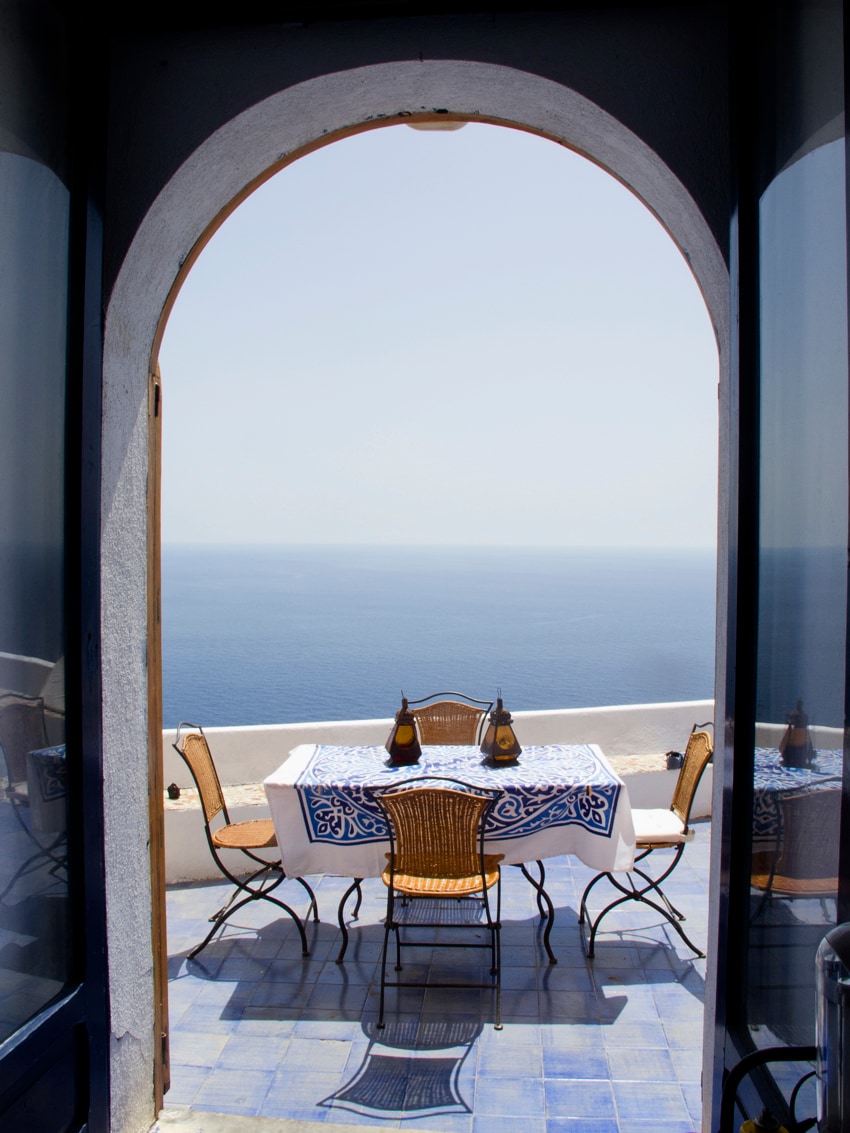 An arched entrance leads to an outdoor terrace with a dining table and chairs. The table is adorned with a blue and white tablecloth. The expansive view showcases the calm sea meeting the horizon under clear skies.