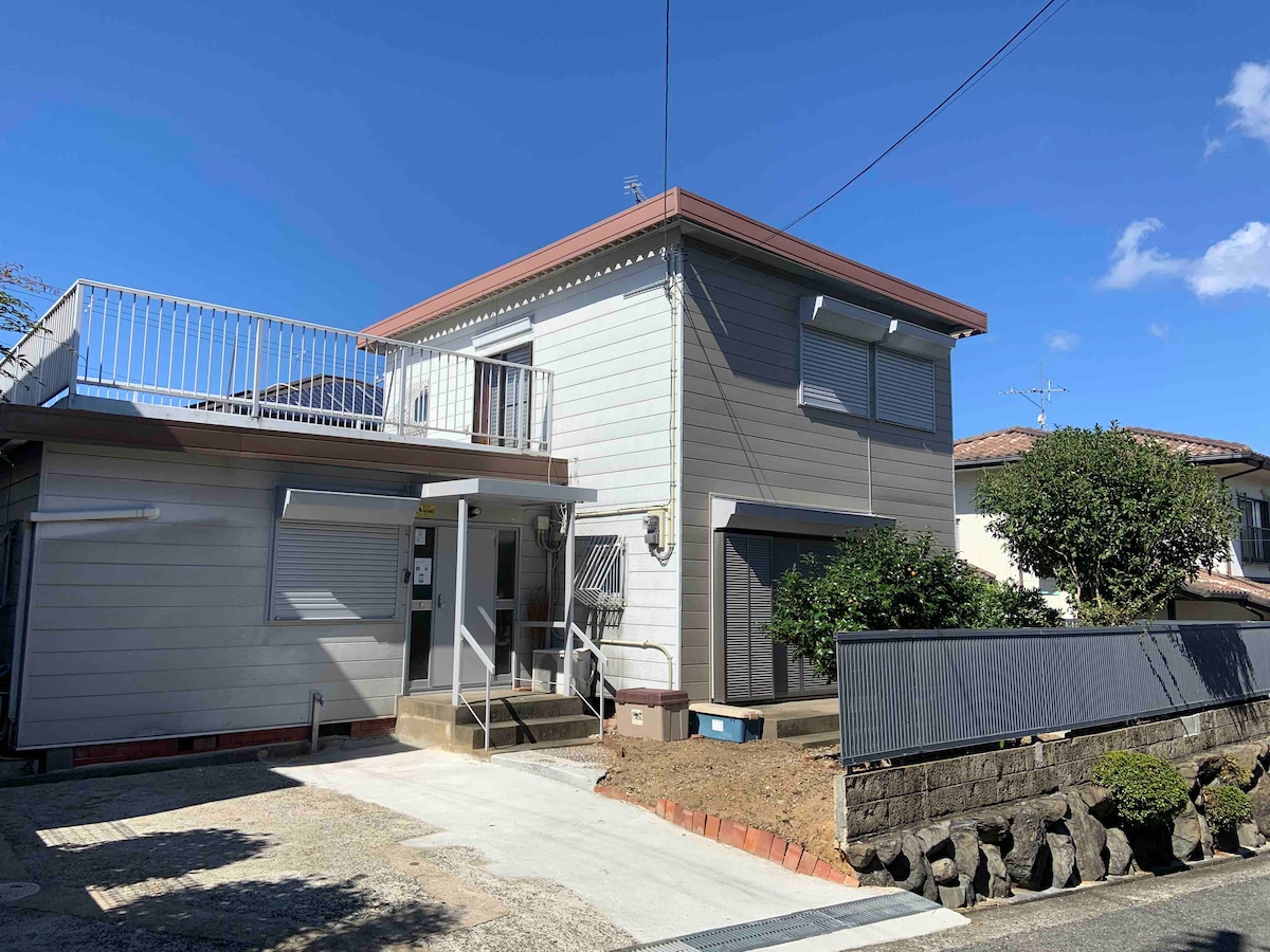 A two-story building is shown, featuring a light-colored exterior and a metal railing on the upper balcony. The entrance is accessible by a pathway leading to the front steps, with a garden area visible on one side. Clear blue skies are seen above.