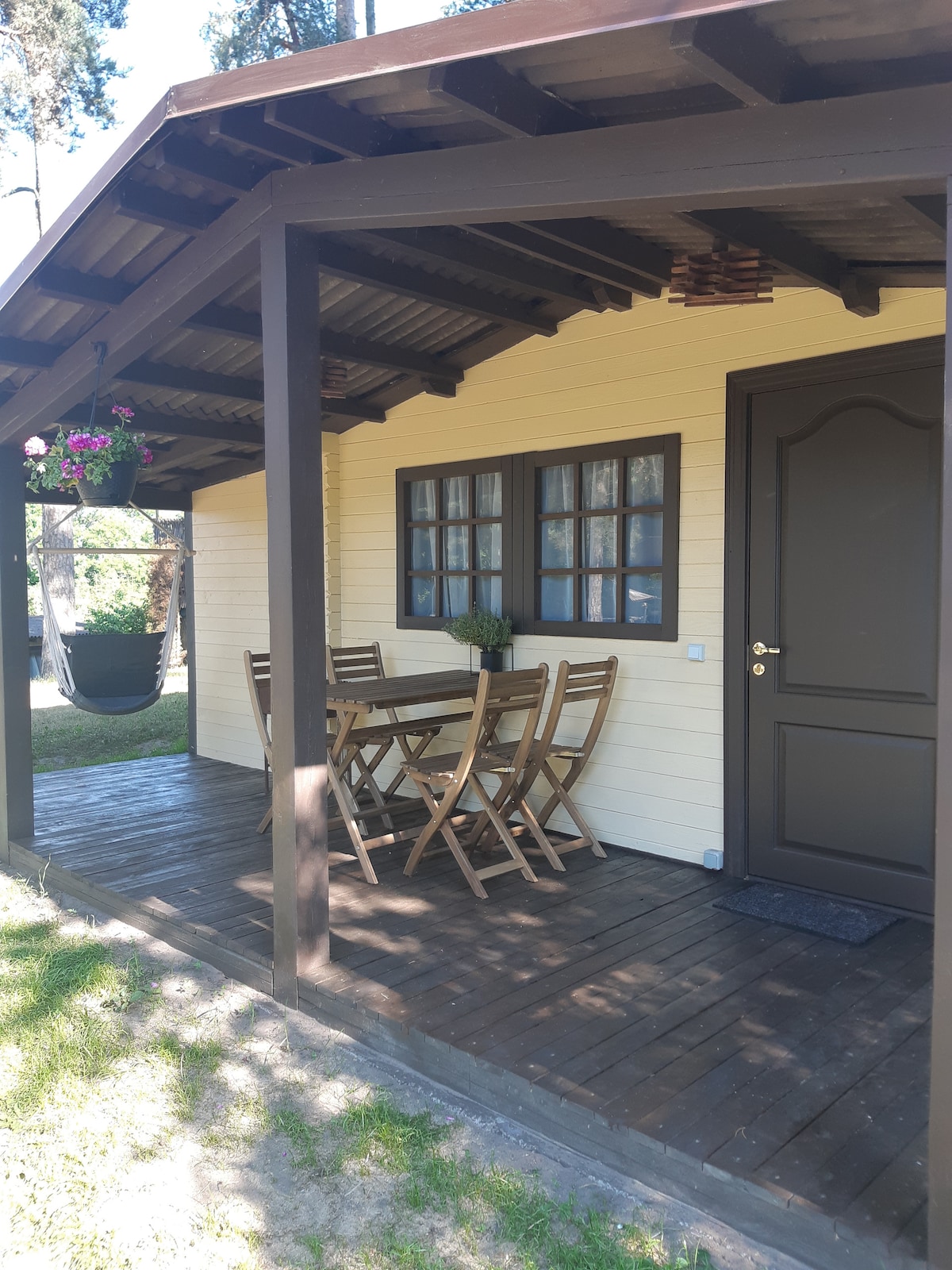 A wooden deck area features a set of six folding chairs surrounding a rectangular table. A welcoming doorway leads into the bungalow, with a light yellow exterior and a series of windows adorned with dark frames. Hanging plants add a touch of greenery.
