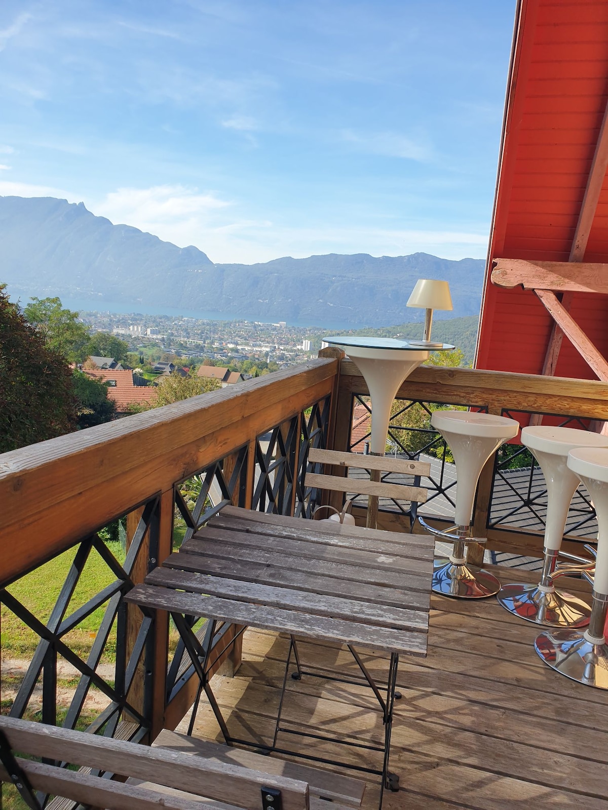 A wooden balcony features a small folding table and several tall stools. The surrounding railing provides a clear view of the mountains and the valley below, under a bright sky illuminated by natural light.