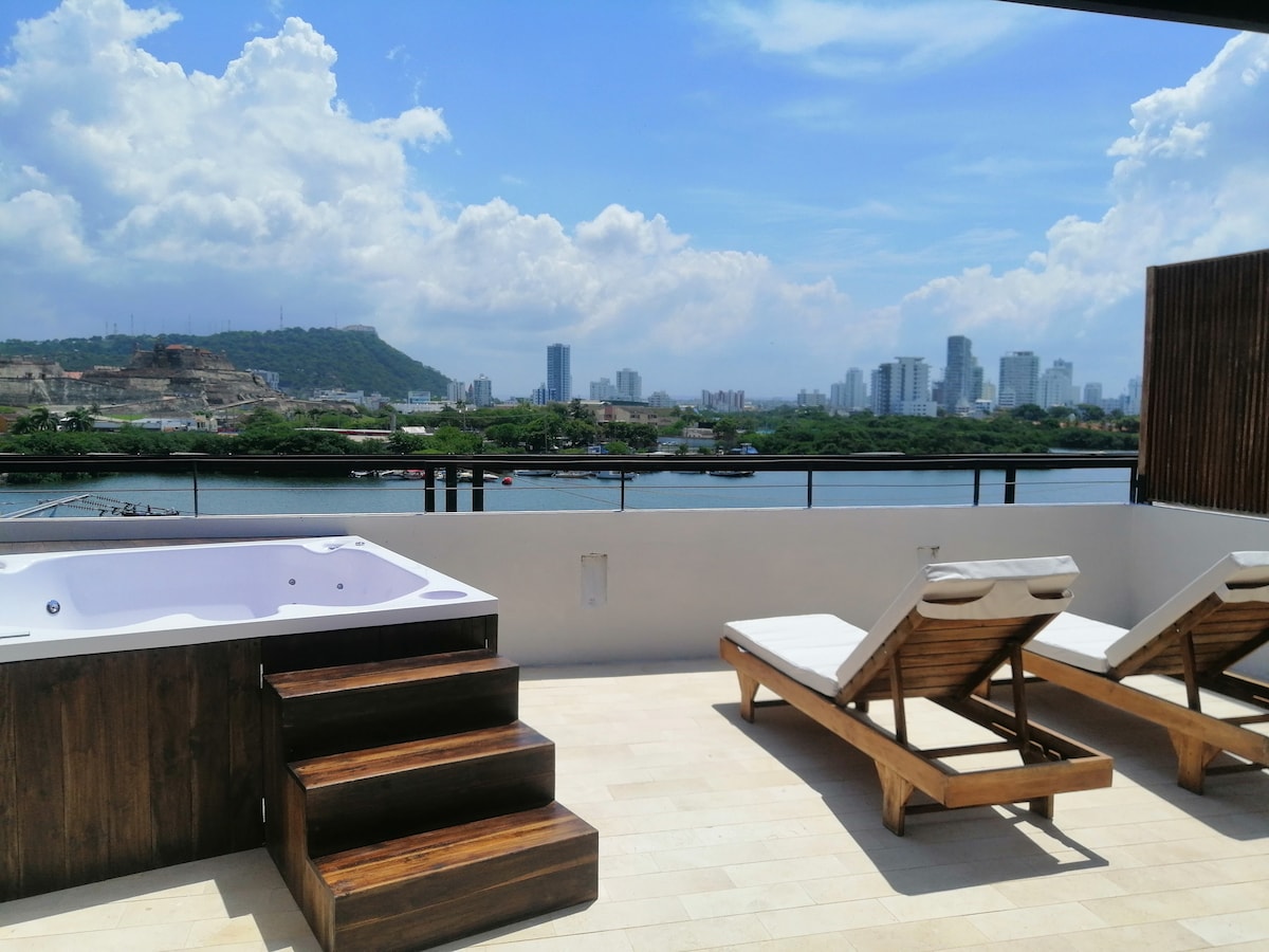 A spacious solarium is presented, featuring a jacuzzi and two wooden lounge chairs. The surface is tiled, leading to a railing with a view overlooking a body of water and the distant skyline, with the Castillo de San Felipe visible in the background.