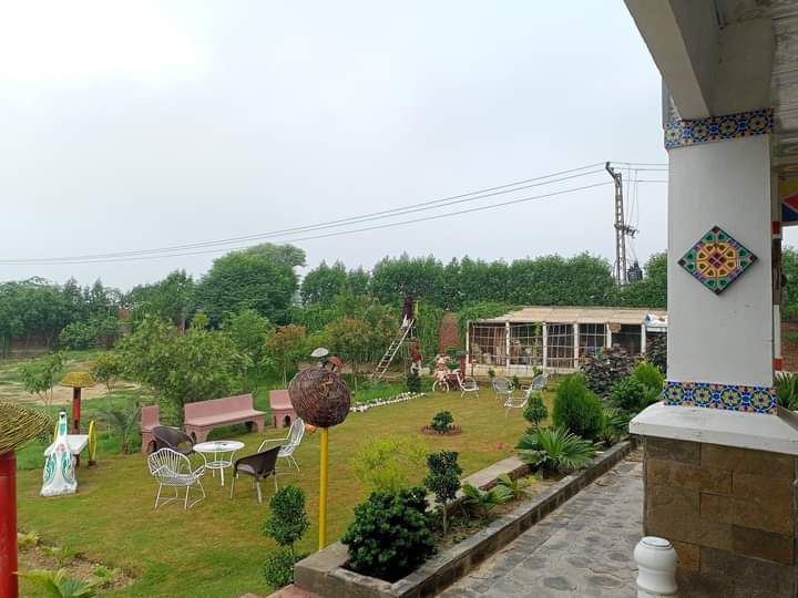 An outdoor space features well-maintained gardens with green grass and various shrubs. Seating arrangements, including white chairs and a pink bench, are positioned for relaxation. A gazebo and swing can be seen in the background, surrounded by trees offering a natural landscape.