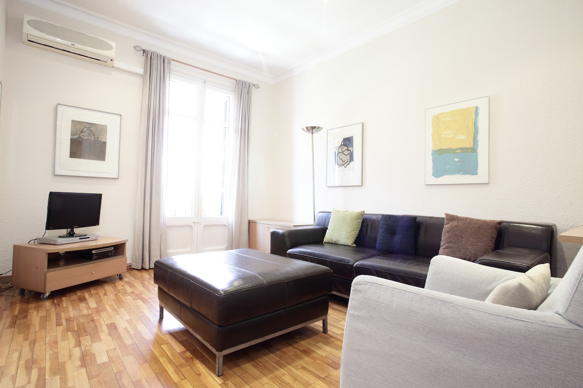 The living room features a comfortable leather sofa and a plush armchair, arranged around a large coffee table. Natural light fills the space through tall windows dressed with light curtains. Artwork decorates the walls, and a television is positioned on a media console.