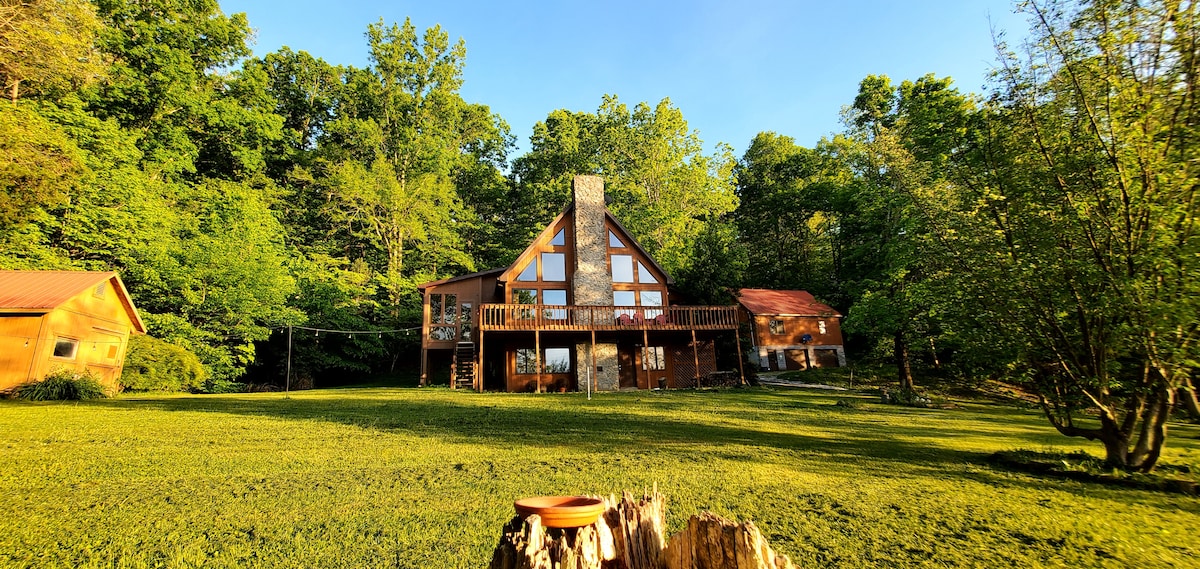The A-frame house features a large central stone chimney and expansive windows, surrounded by lush green foliage. A wooden deck extends across the front, providing a welcoming space to enjoy the outdoors. The well-maintained lawn offers an inviting atmosphere for relaxation.
