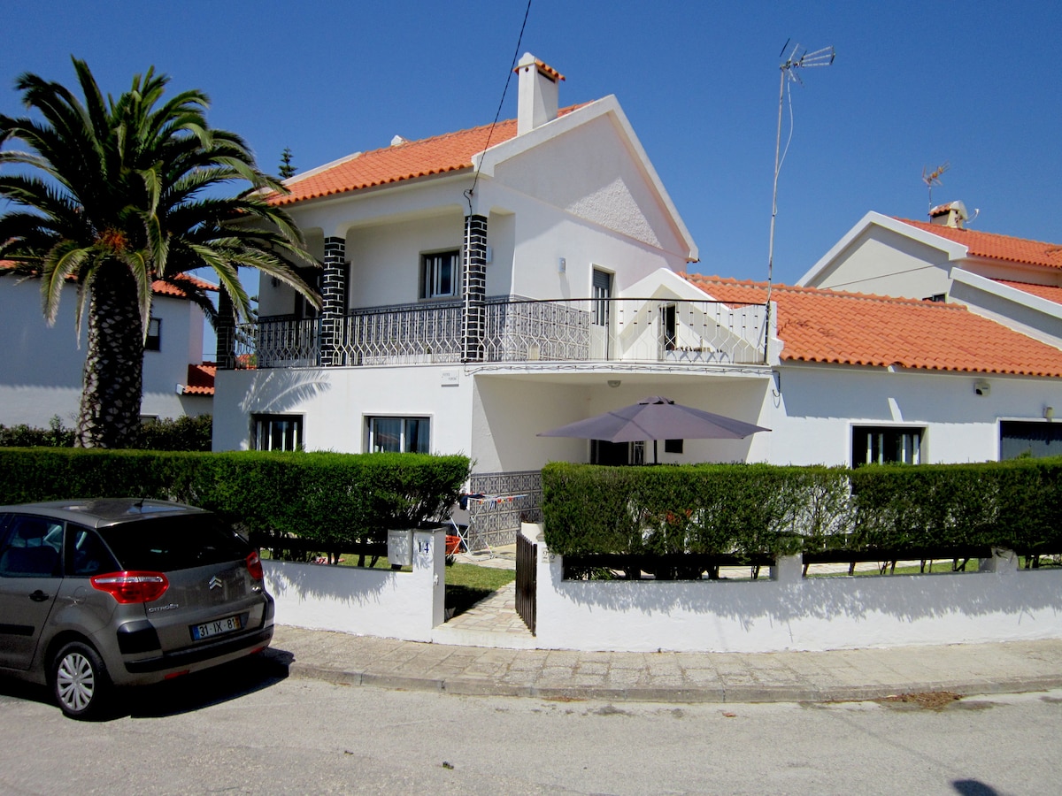 A two-story house is shown with a light exterior and a red-tiled roof. Lush green hedges line the property, and a car is parked in front. A shaded area is provided by a large umbrella near the entrance, complemented by palm trees in the background.