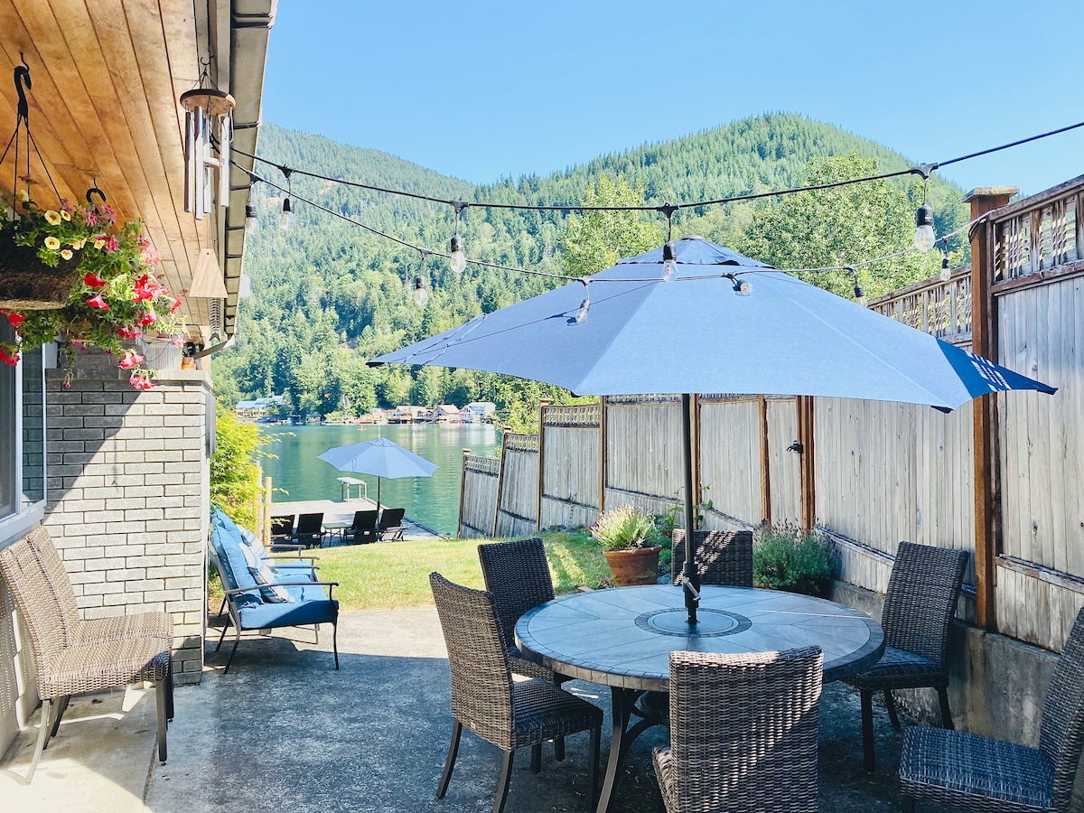 A lakeside patio features a round table surrounded by six woven chairs, shaded by a large blue umbrella. String lights are draped overhead, and a view of the lake and surrounding greenery is visible, creating an inviting outdoor space for relaxation.