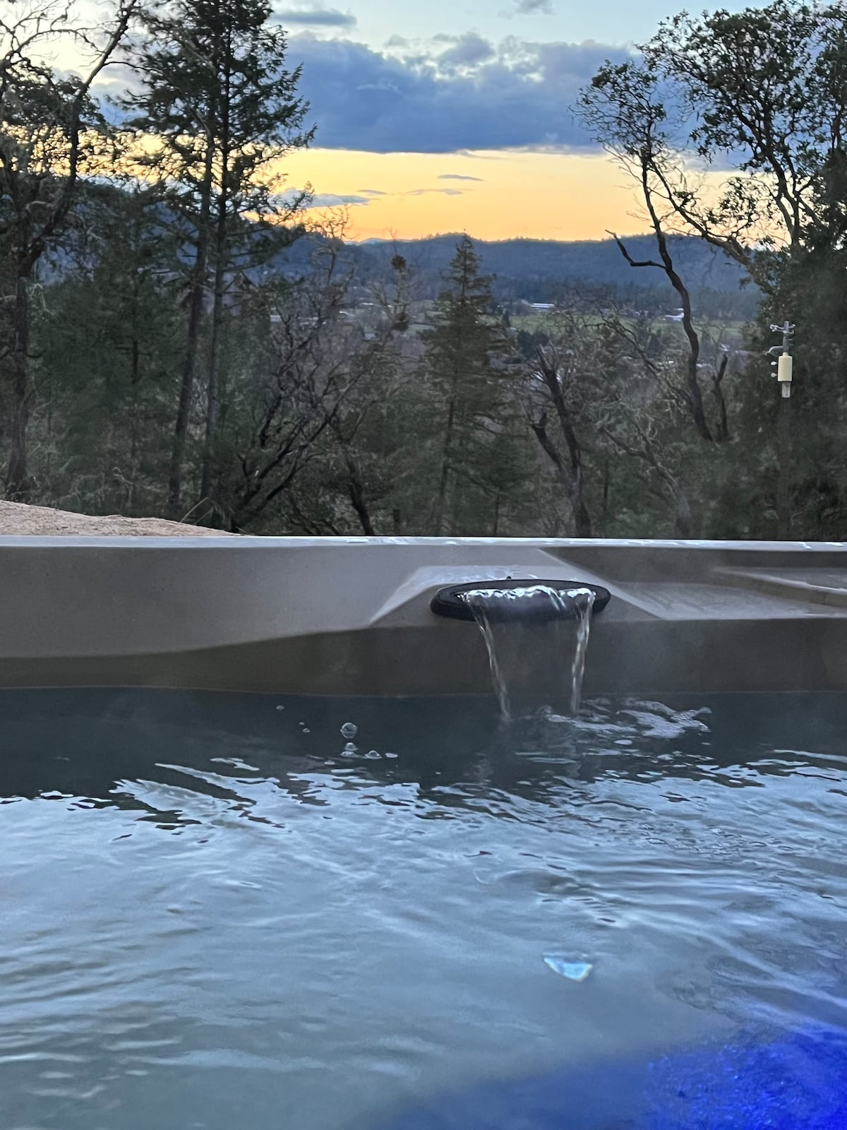 A hot tub is positioned with a scenic view of the valley and trees in the background. Water cascades gently from the edge of the tub, reflecting the soft colors of the sunset in the sky.