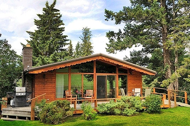 A charming cabin is nestled among tall trees, featuring a wooden deck and large glass doors that provide ample natural light. A grill is placed on the deck for outdoor cooking, with comfortable chairs inviting relaxation in the surrounding greenery.