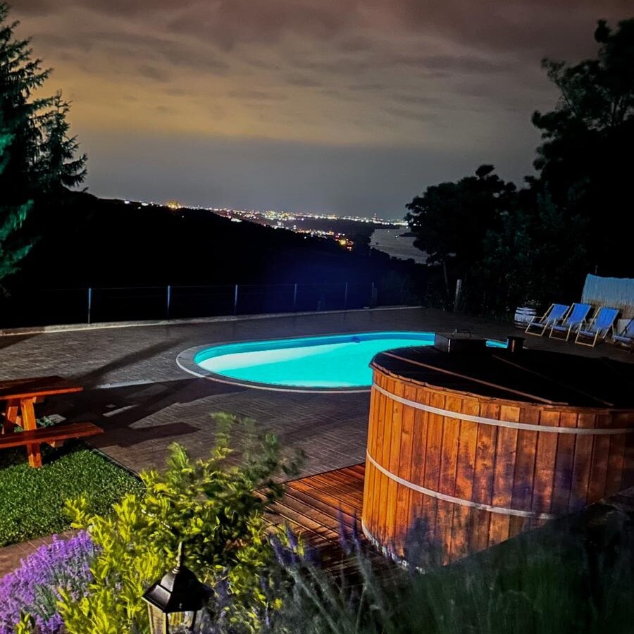 The outdoor area features a private pool illuminated in soft blue light, surrounded by wooden seating and a hot tub. A picnic table and lounge chairs offer a comfortable space to relax, with a view of the distant city lights against the night sky.