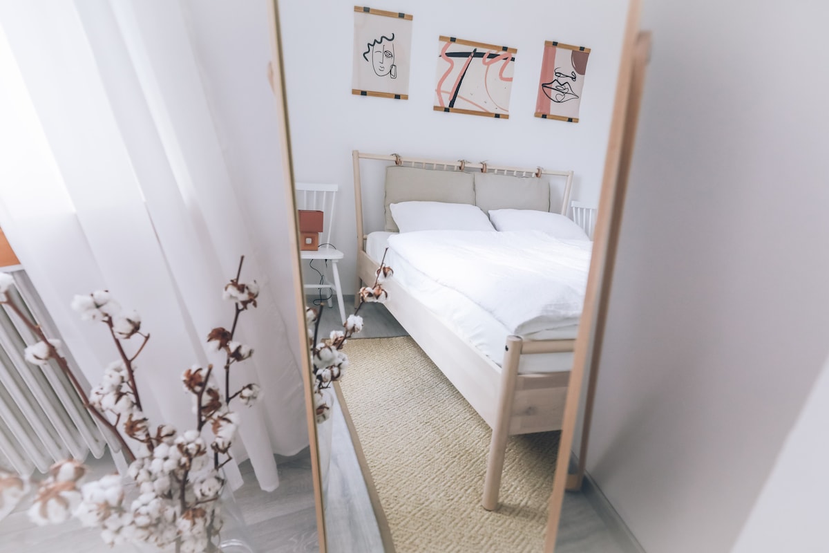 A bright bedroom features a wide bed with white bedding, reflected in a full-length mirror. Soft, neutral tones are complemented by abstract wall art. A nearby bedside table holds a small lamp, while a decorative plant adds a natural touch to the space.