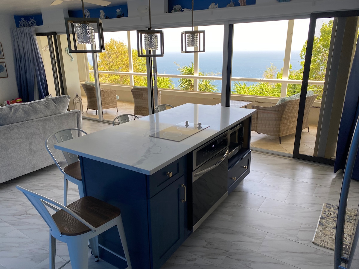 The kitchen area features a central island with a countertop stove and modern appliances. Barstools are positioned around the island, providing additional seating. Floor-to-ceiling glass doors lead to a balcony, offering an expansive ocean view complemented by greenery.