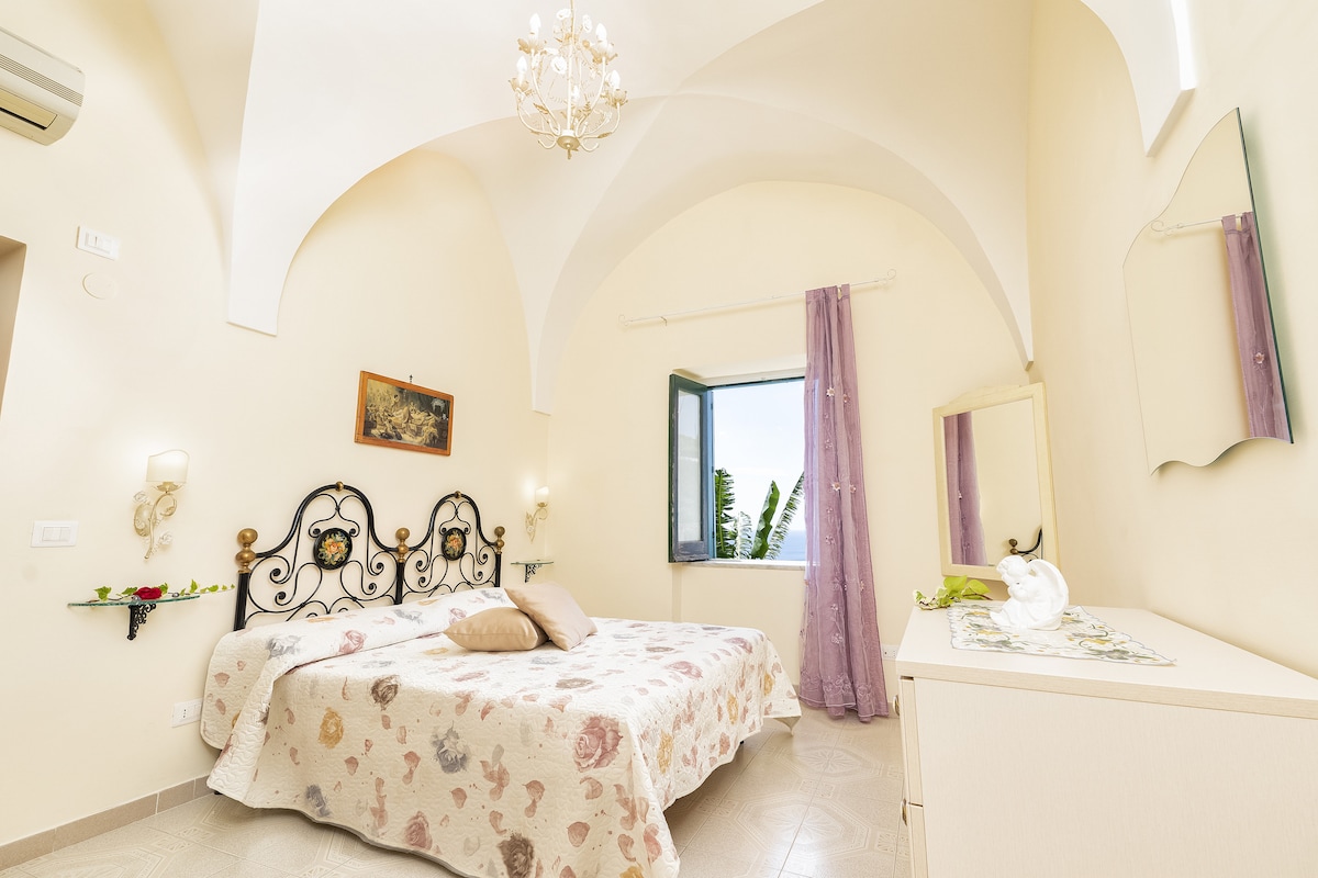 A spacious bedroom is adorned with arched ceilings and soft, neutral-toned walls. A large bed is positioned centrally, complemented by decorative headboards. Natural light enters through an open window, illuminating a dresser and a vase placed elegantly on a table.
