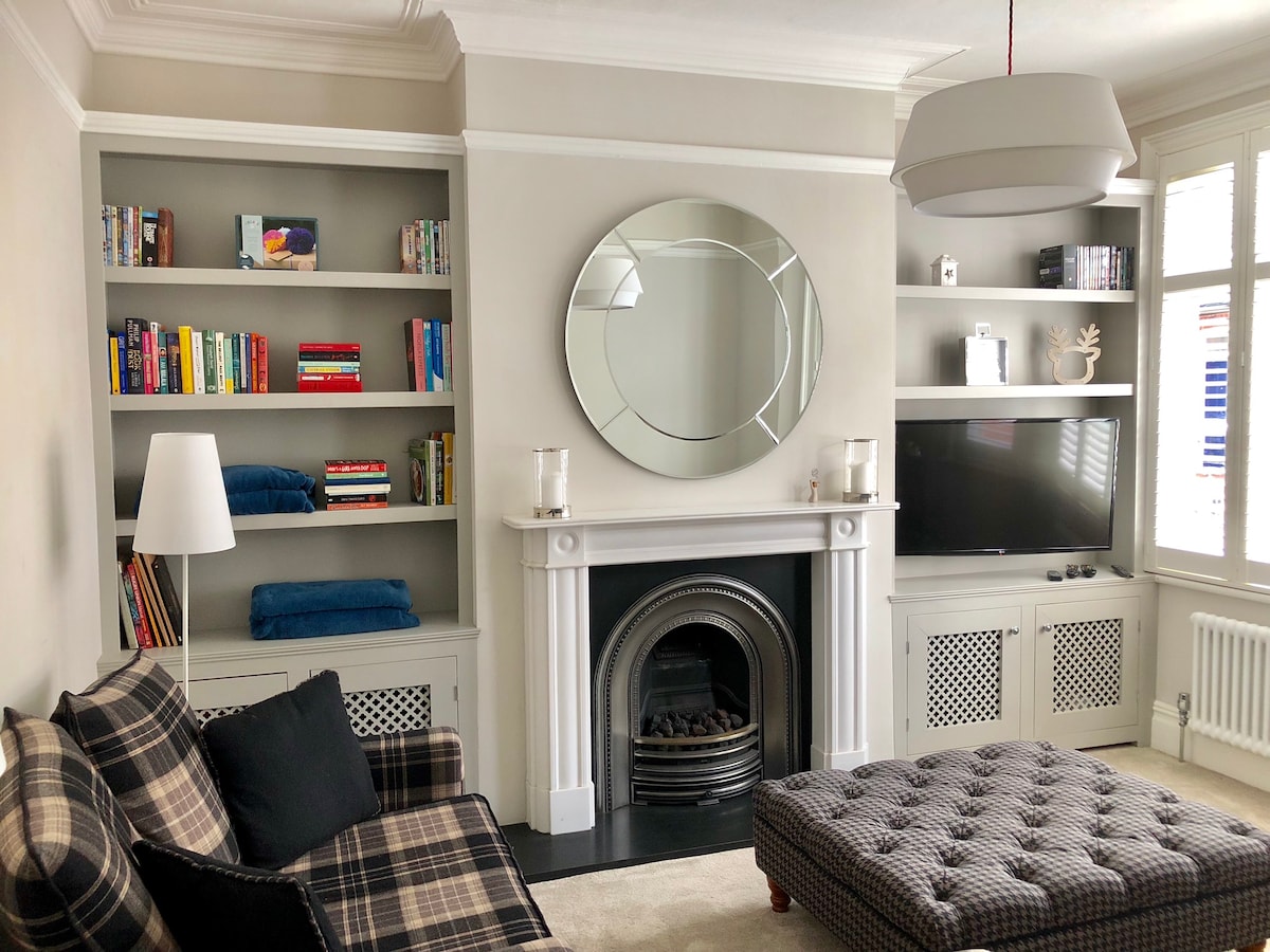 A comfortable sitting room is presented, featuring a three-seater sofa and two-seater sofa, complemented by a large ottoman. A classic marble and granite fireplace with a remote-controlled fire is at the center. The room includes built-in shelves displaying books, and a TV is mounted on the wall.