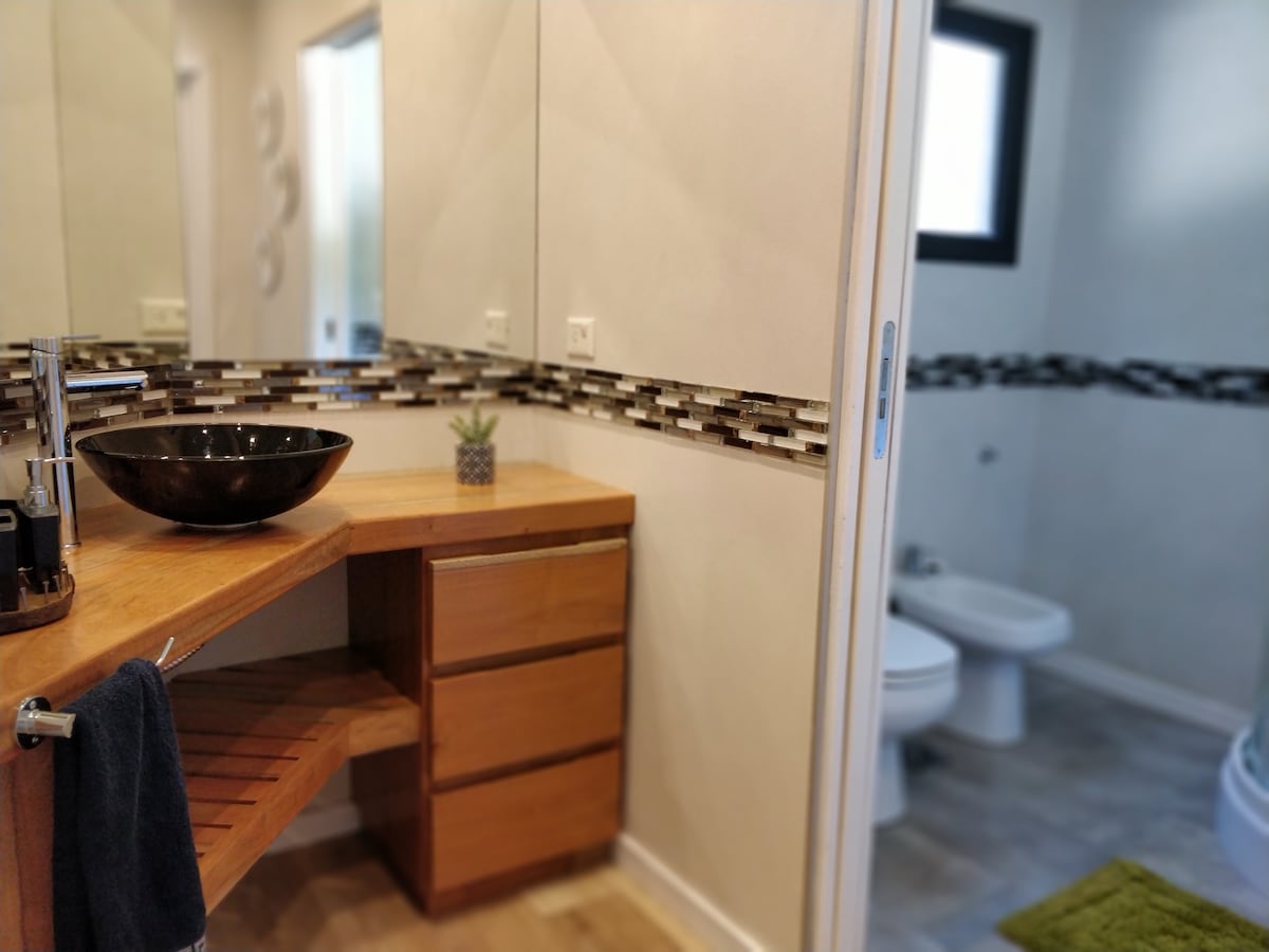 The bathroom features a modern wooden vanity with a dark bowl sink, complemented by a mix of tiled accents along the wall. Soft natural light enters from a window, illuminating the spacious layout. To the side, a glimpse of the restroom area can be seen.