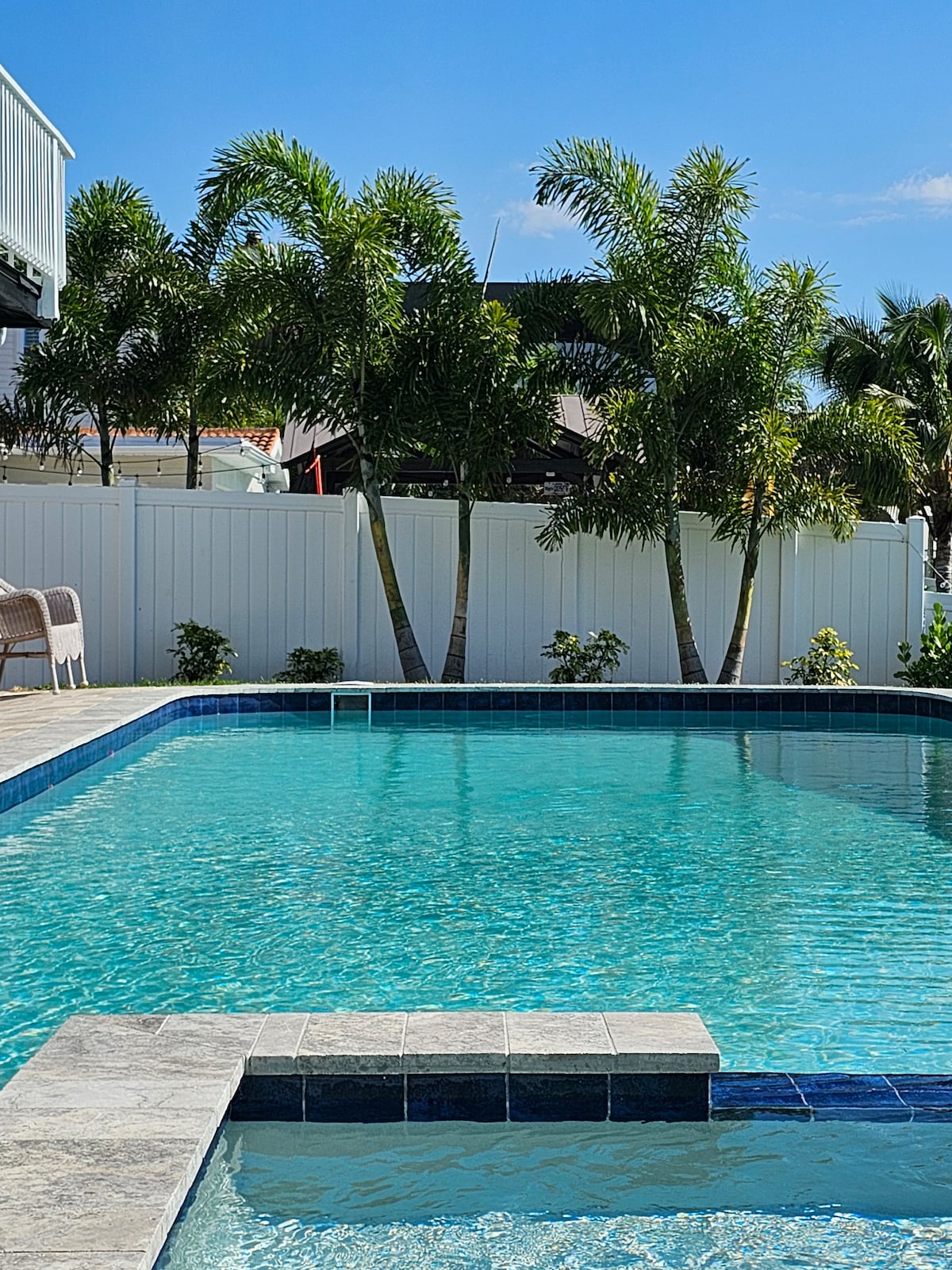 A large lap pool with clear blue water is surrounded by a paved area and lush palm trees. A white fence provides privacy, while bright sunlight enhances the inviting atmosphere. The pool features a spa area with steps for easy access.