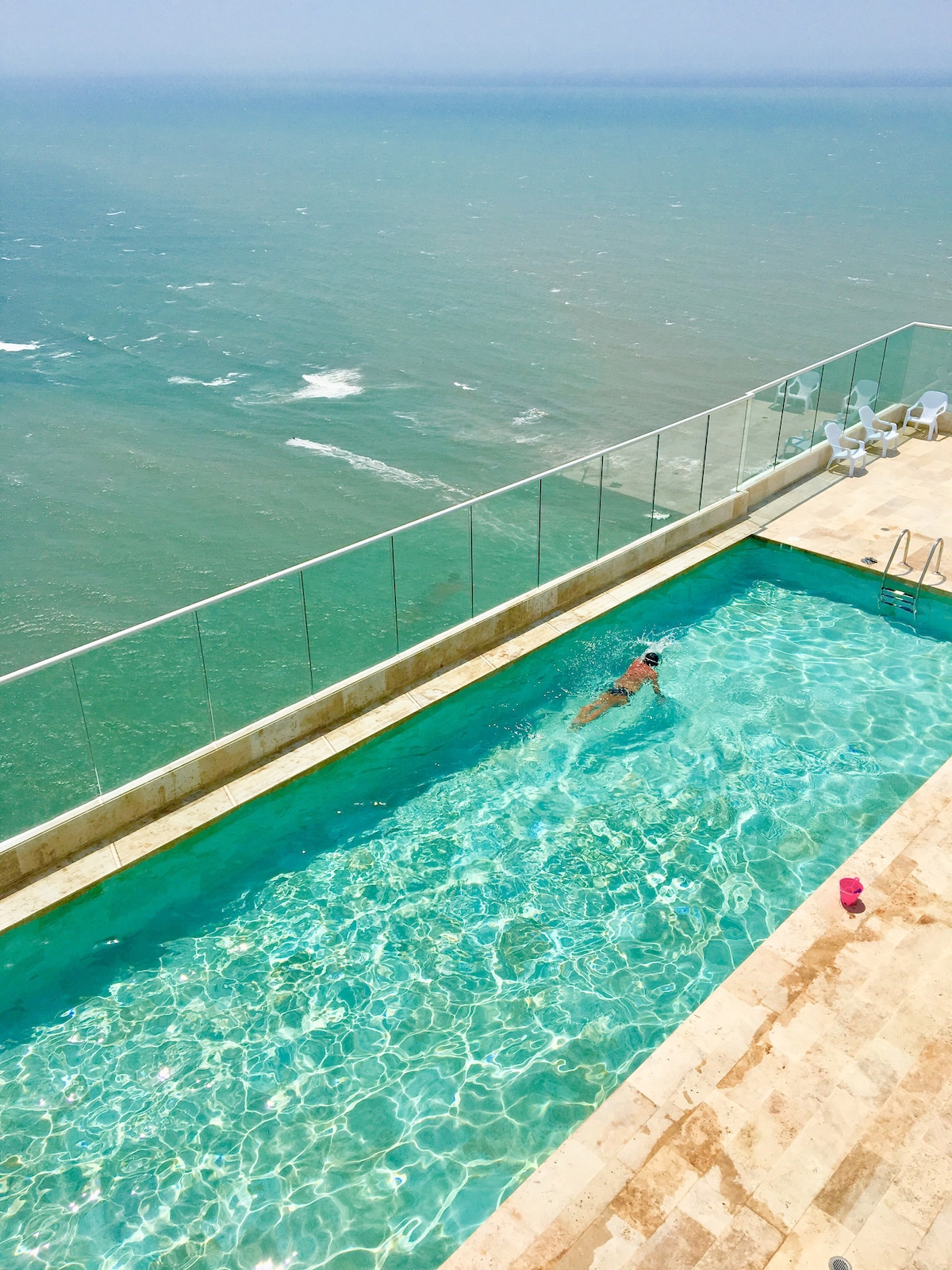 A rooftop swimming pool overlooks the ocean, featuring clear turquoise water. A swimmer can be seen enjoying the space, while white lounge chairs line the edge of the pool area, providing a place to relax and take in the expansive sea views.