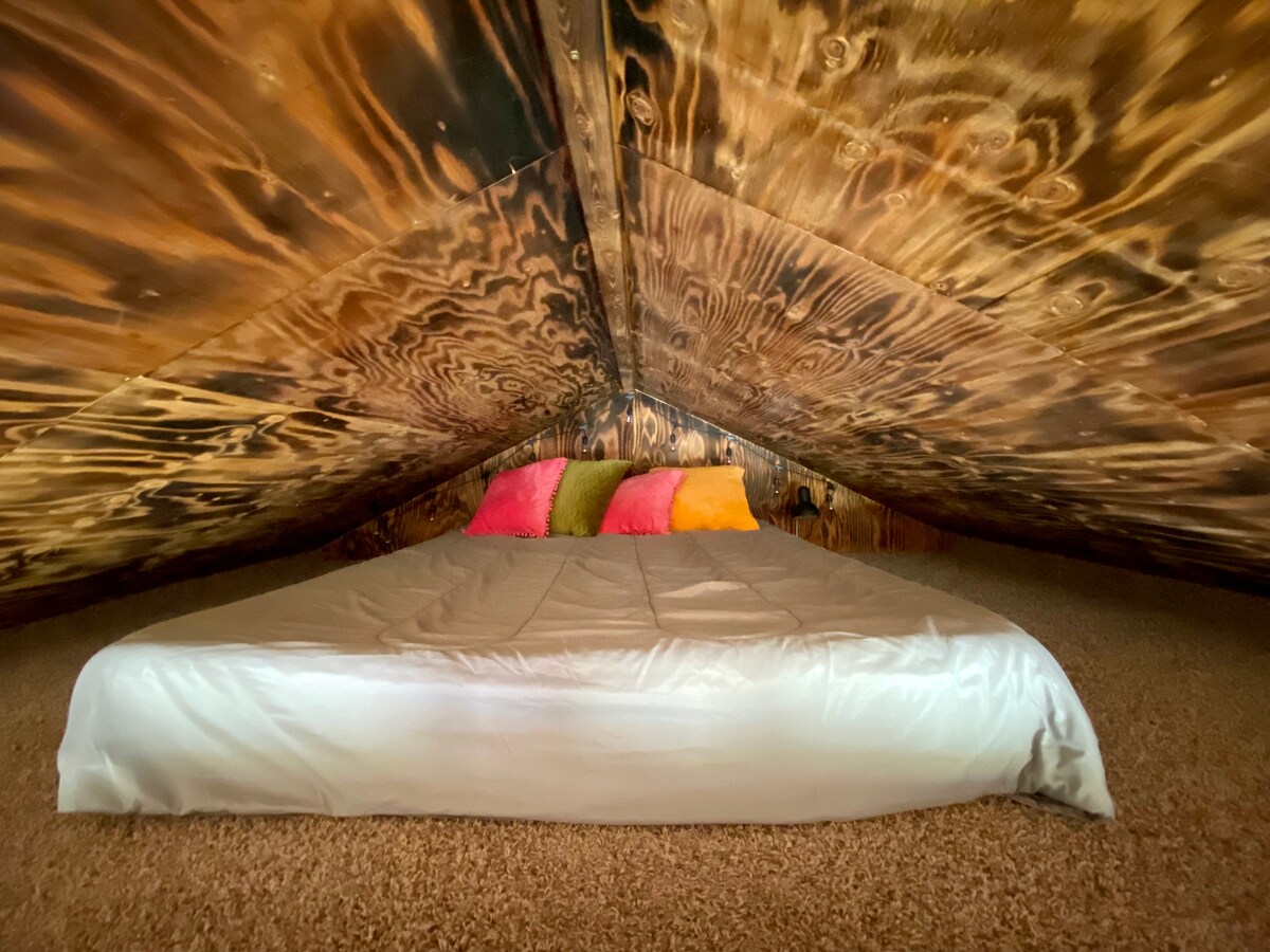 An elevated sleeping area is presented under a wooden, sloped ceiling with distinctive grain patterns. A spacious bed is positioned centrally, adorned with a neutral-colored bedding and colorful accent pillows in shades of pink, orange, and green, set against a warm, rustic backdrop.