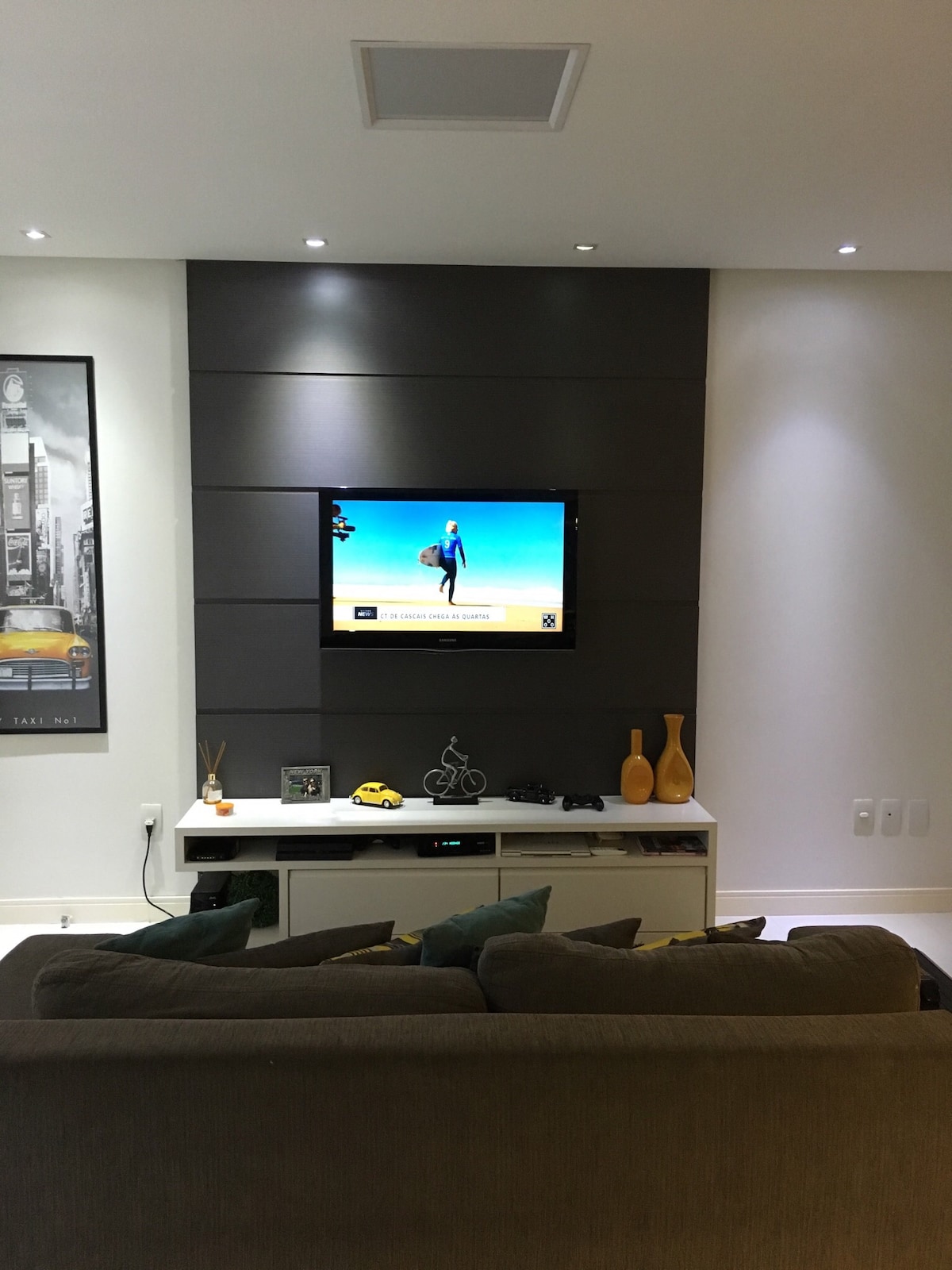 A modern living room is showcased with a low-profile couch adorned with green cushions. A sleek entertainment unit is positioned beneath a mounted flat-screen television. Ambient lighting highlights the textured wall behind, while decorative vases add a touch of elegance to the space.