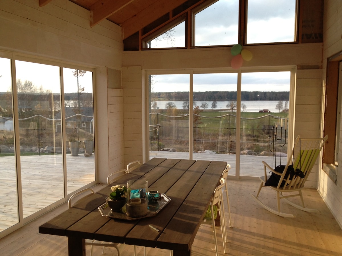 A spacious dining area features large glass doors that provide a clear view of the water and surrounding landscape. A wooden table is set with decorative items, while light-colored chairs surround it. Natural light floods the space, highlighting the wooden accents and open layout.
