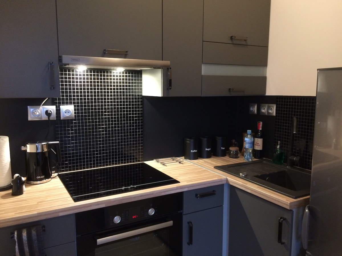 A modern kitchen is shown with dark cabinetry and a wooden countertop. An electric stovetop and oven are positioned below a sleek hood, while a sink is located beside the stylish black tiled backsplash. Kitchen appliances are neatly organized, enhancing functionality.