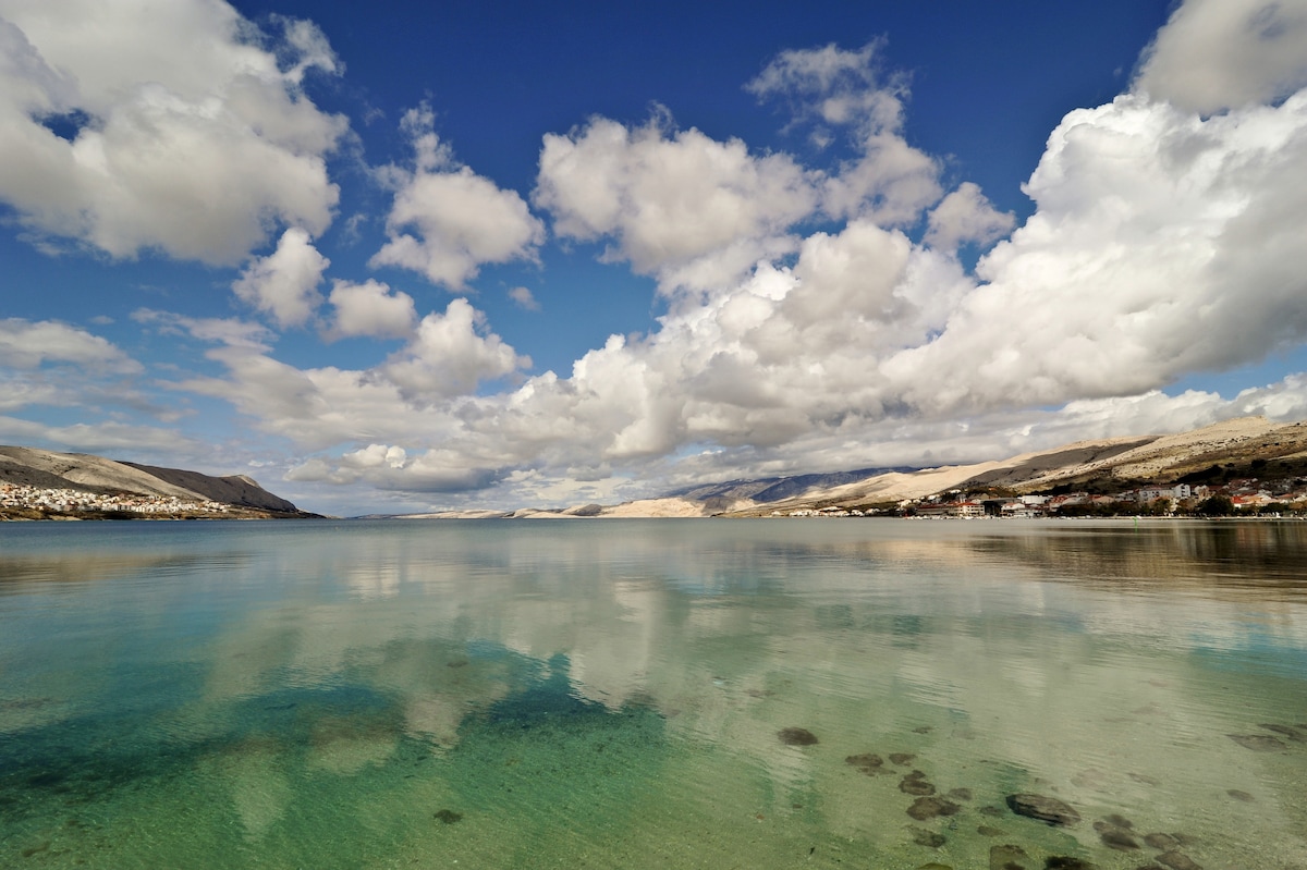 A serene coastal view features calm waters reflecting a dramatic sky adorned with fluffy clouds. The shoreline is dotted with faint outlines of the town and distant hills, emphasizing the tranquil connection between land and sea.