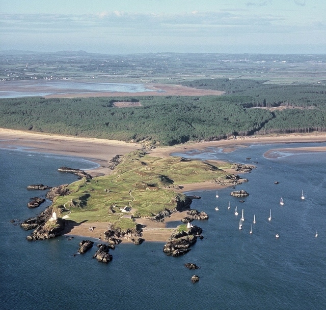 Aerial view of a coastal landscape featuring an island with lush green hills and rocky formations. A series of sailboats can be seen in the surrounding water, while sandy beaches extend along the shoreline and a forested area is visible in the background.