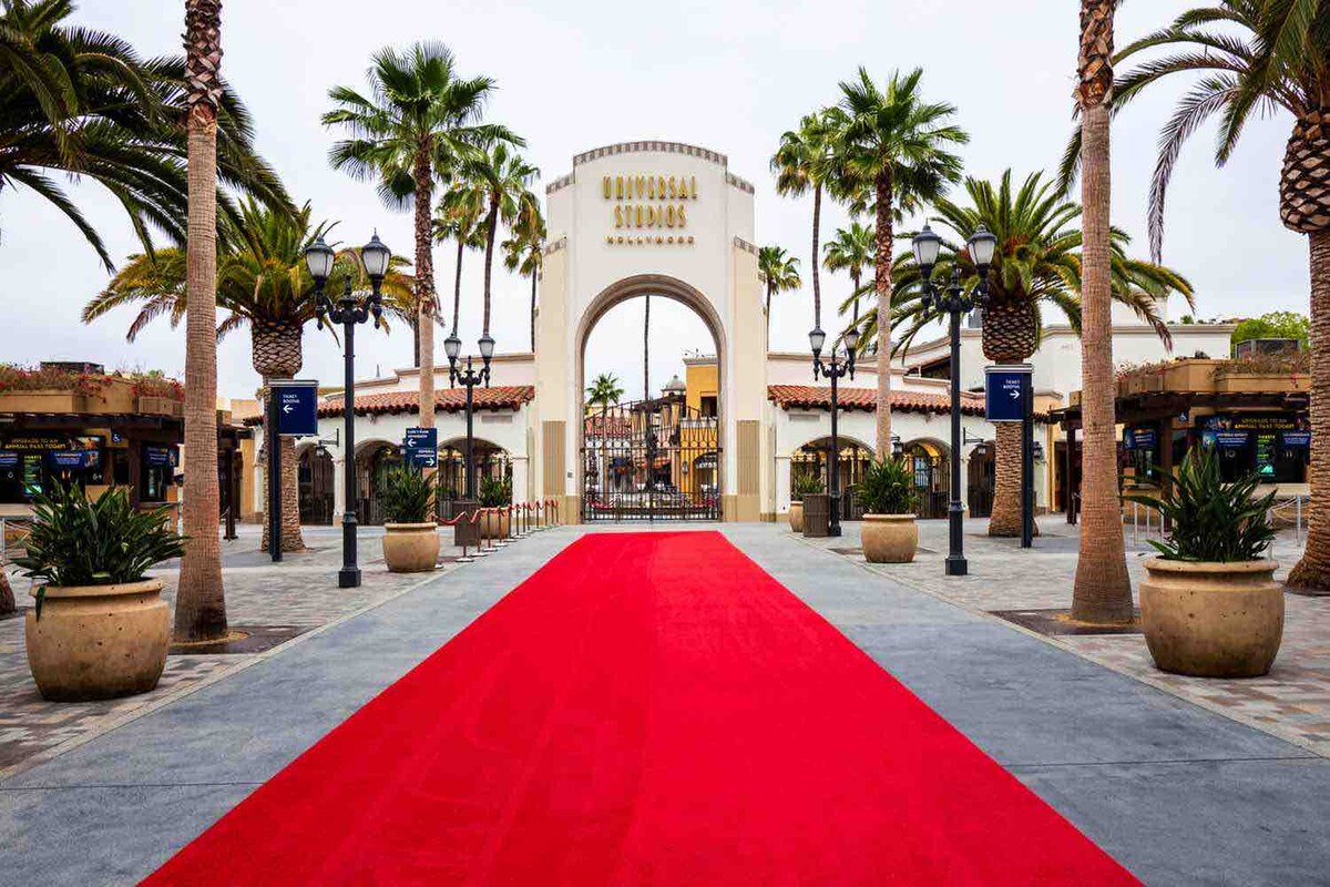 The entrance to Universal Studios is showcased, featuring a prominent red carpet leading toward the iconic archway. Lush palm trees frame the scene, and decorative planters add greenery to the paved pathway, inviting visitors into the vibrant theme park atmosphere.