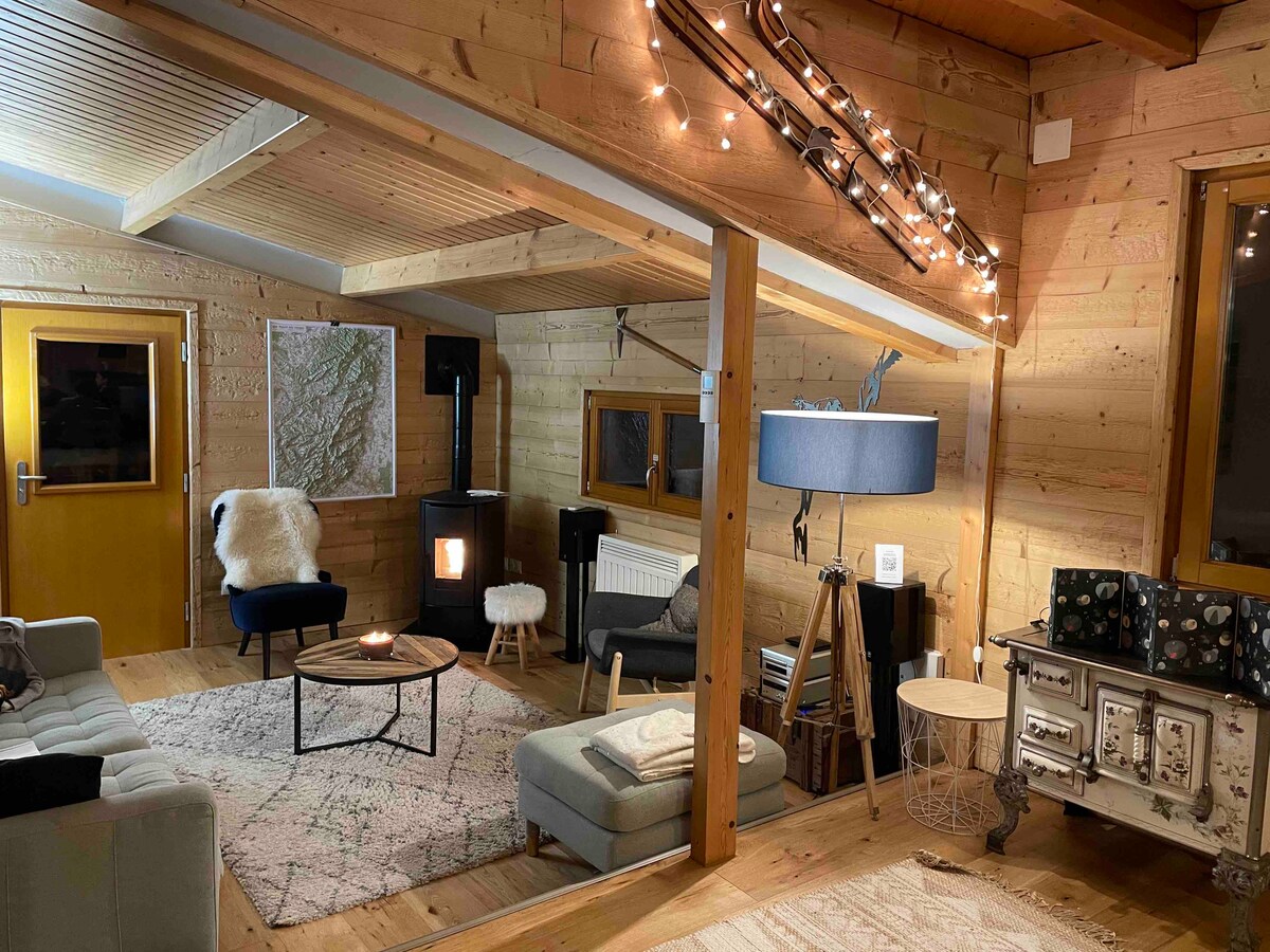 A warm living area is characterized by wooden walls and a high ceiling. Soft seating options surround a circular table, with a pellet stove providing heating. Ambient lighting is created by string lights adorning the ceiling. A floor lamp and a wall map are visible, enhancing the space.