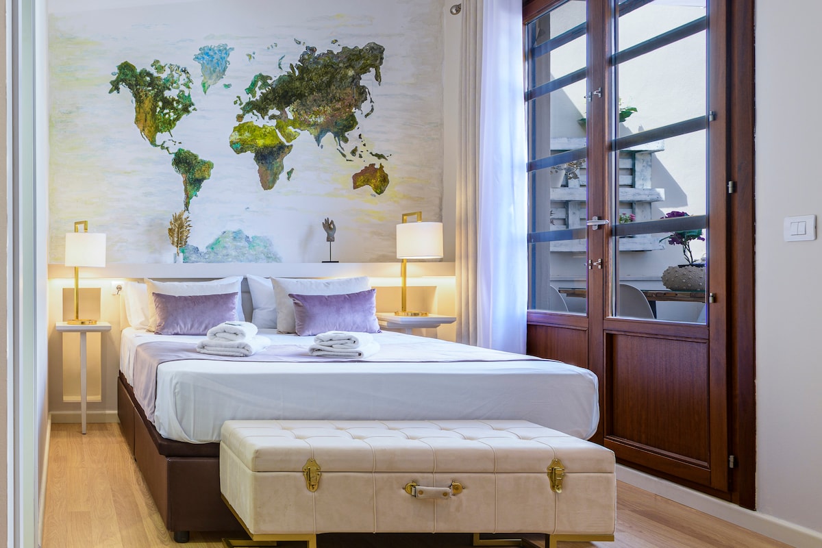 A sleeping area features a spacious bed with neatly arranged linens. A large world map mural adorns the wall, complemented by soft lighting from two bedside lamps. Light streams through large glass doors, revealing a view of a balcony with greenery.